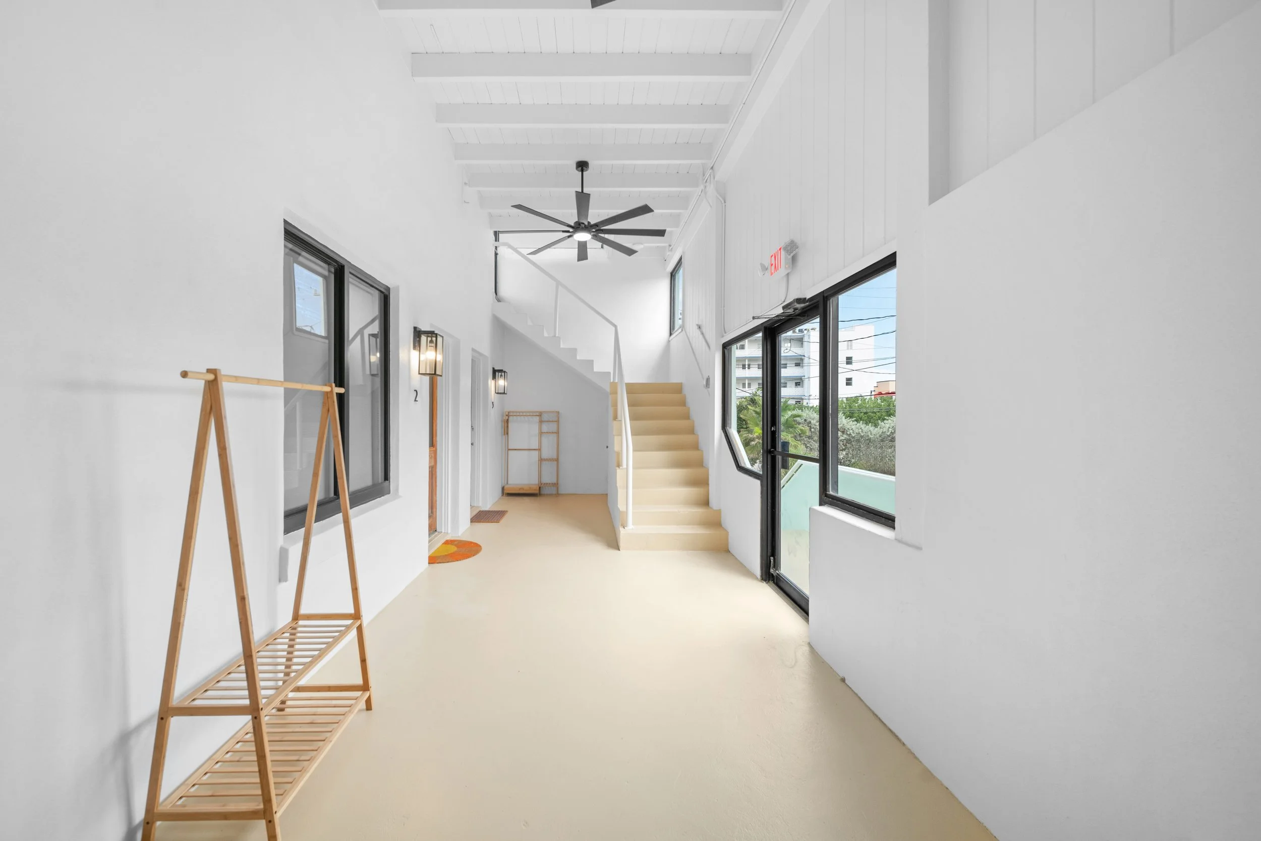 Bright, modern hallway with white walls, black-framed windows, staircase, ceiling fan, and wooden furniture.