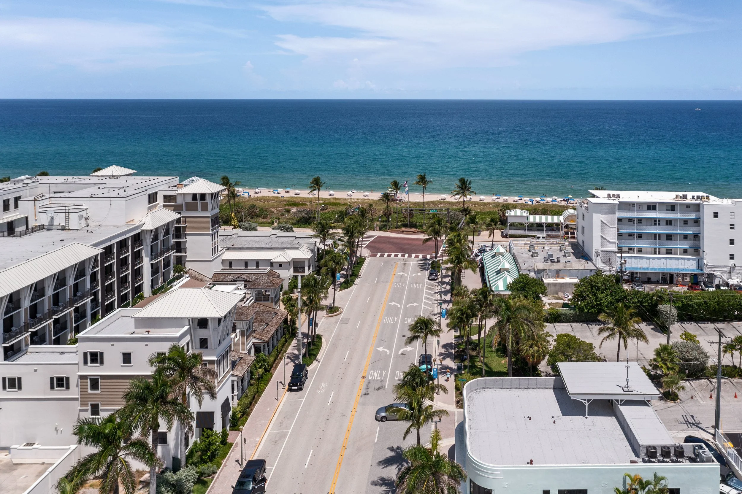 Aerial view of a coastal city street leading to a beach with umbrellas and palm trees, with buildings on either side and the ocean in the background.