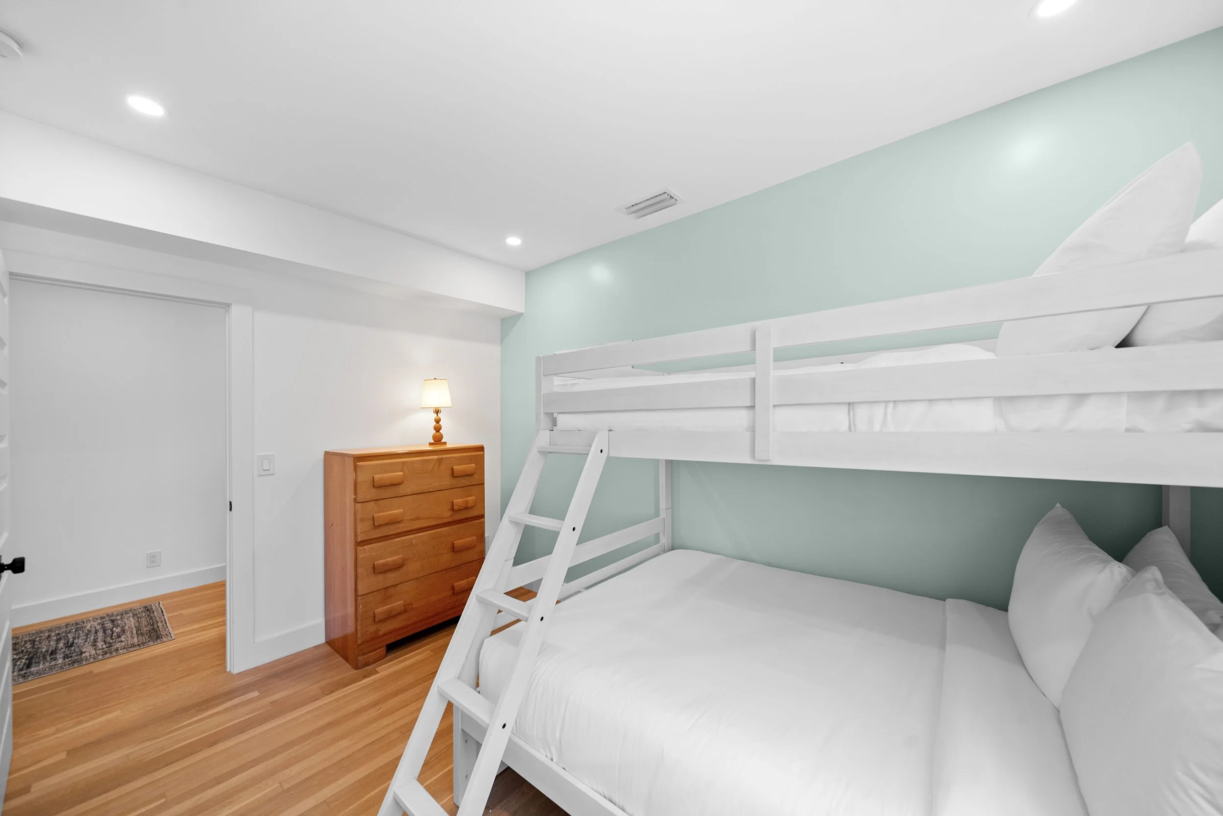 Room with white bunk beds, a wooden dresser with a lamp, and hardwood floors.