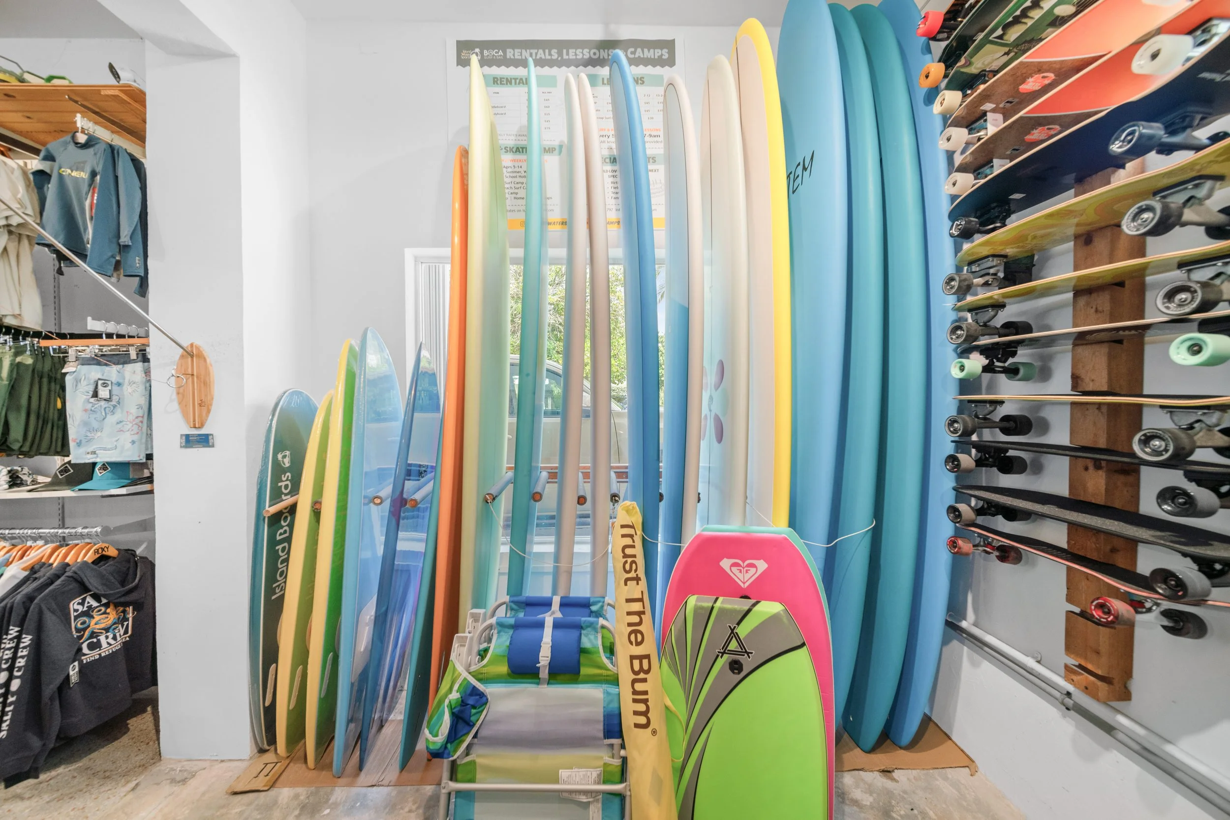 Display of surfboards arranged vertically on a wall stand inside a retail store, with skateboard decks on a rack to the right and clothing on shelves to the left.