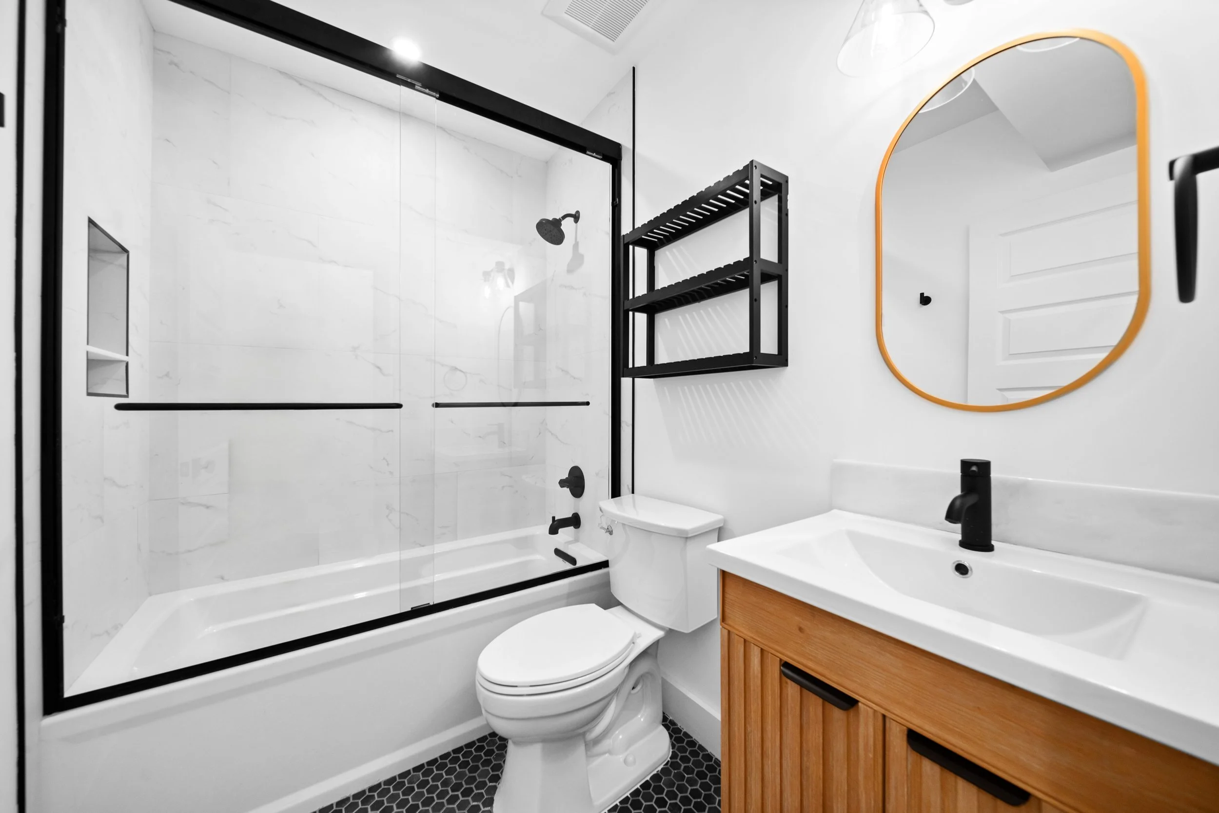 Modern bathroom with a glass-enclosed shower, white toilet, wooden vanity with a white sink, black faucet, black wall shelf, and an orange-framed mirror.
