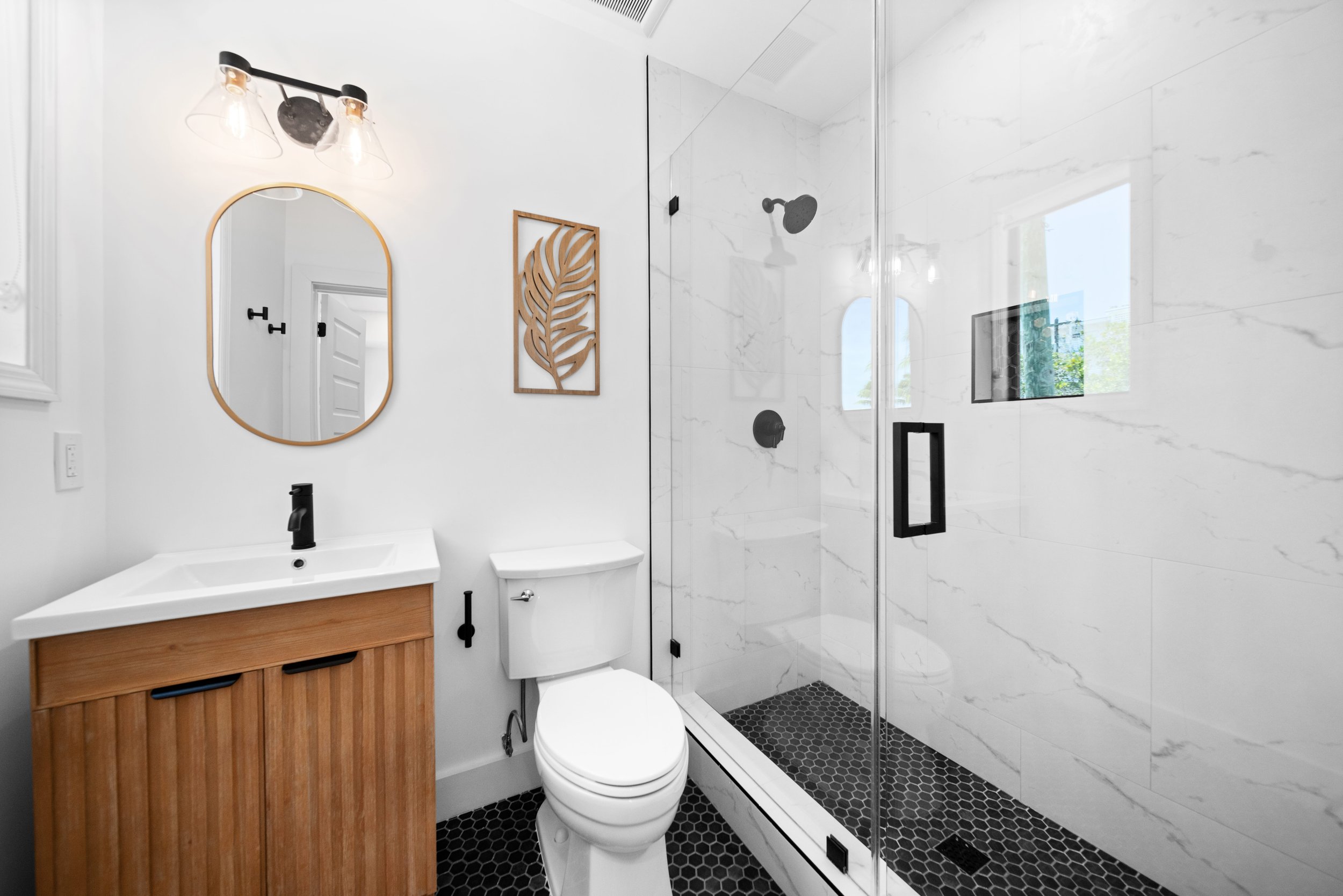 Modern bathroom with white walls, black fixtures, a wooden vanity with a white sink, a toilet, a walk-in shower with a glass door, black hexagonal tiles, and natural light from a small window.