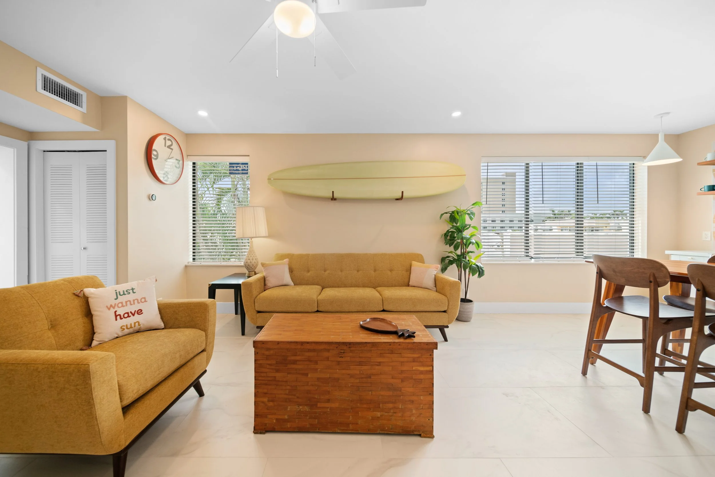 A living room with two mustard-colored couches, a wooden coffee table, a surfboard on the wall, a potted plant, a side table with a lamp, and windows with blinds allowing natural light.