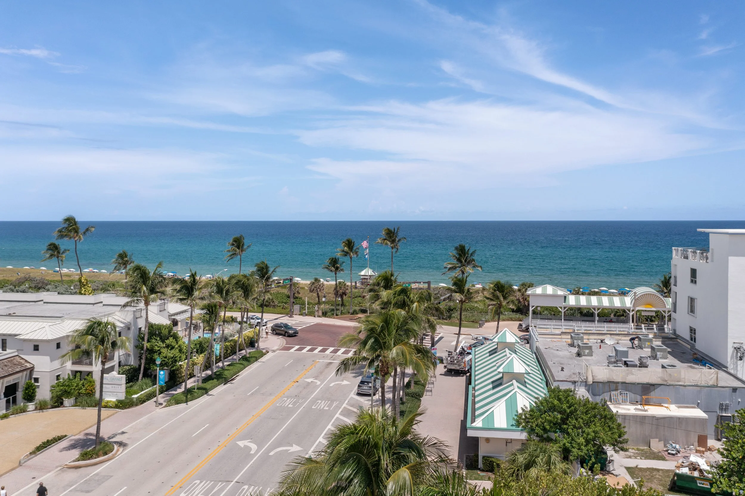 A beachside view with a road lined with palm trees, leading to the ocean. There are beach chairs, a gazebo, and a white building, with blue skies and some clouds overhead.