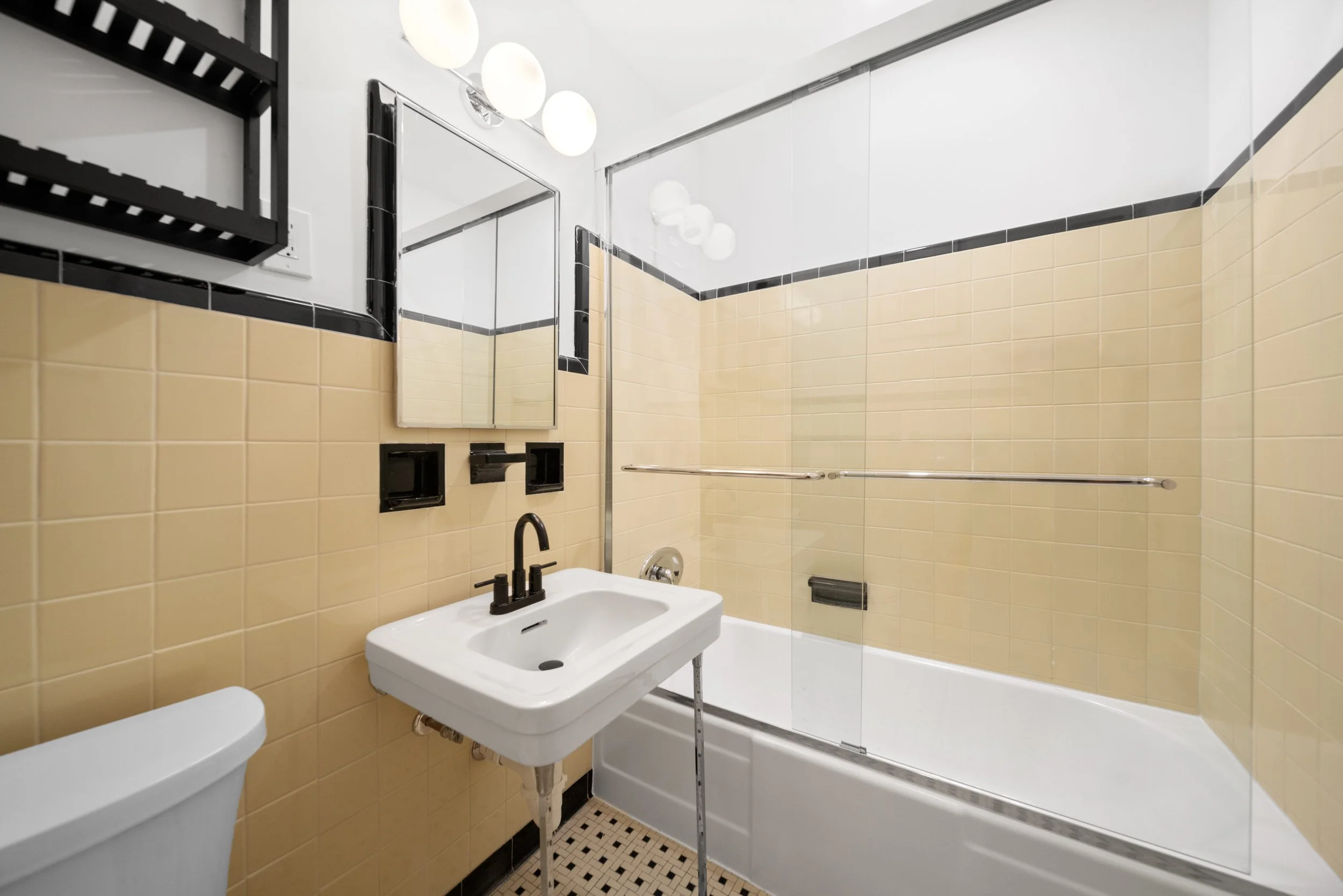 Bathroom with beige tiled walls, a white sink with black fixtures, a mirror, a toilet, and a bathtub with a glass shower partition and black accents.