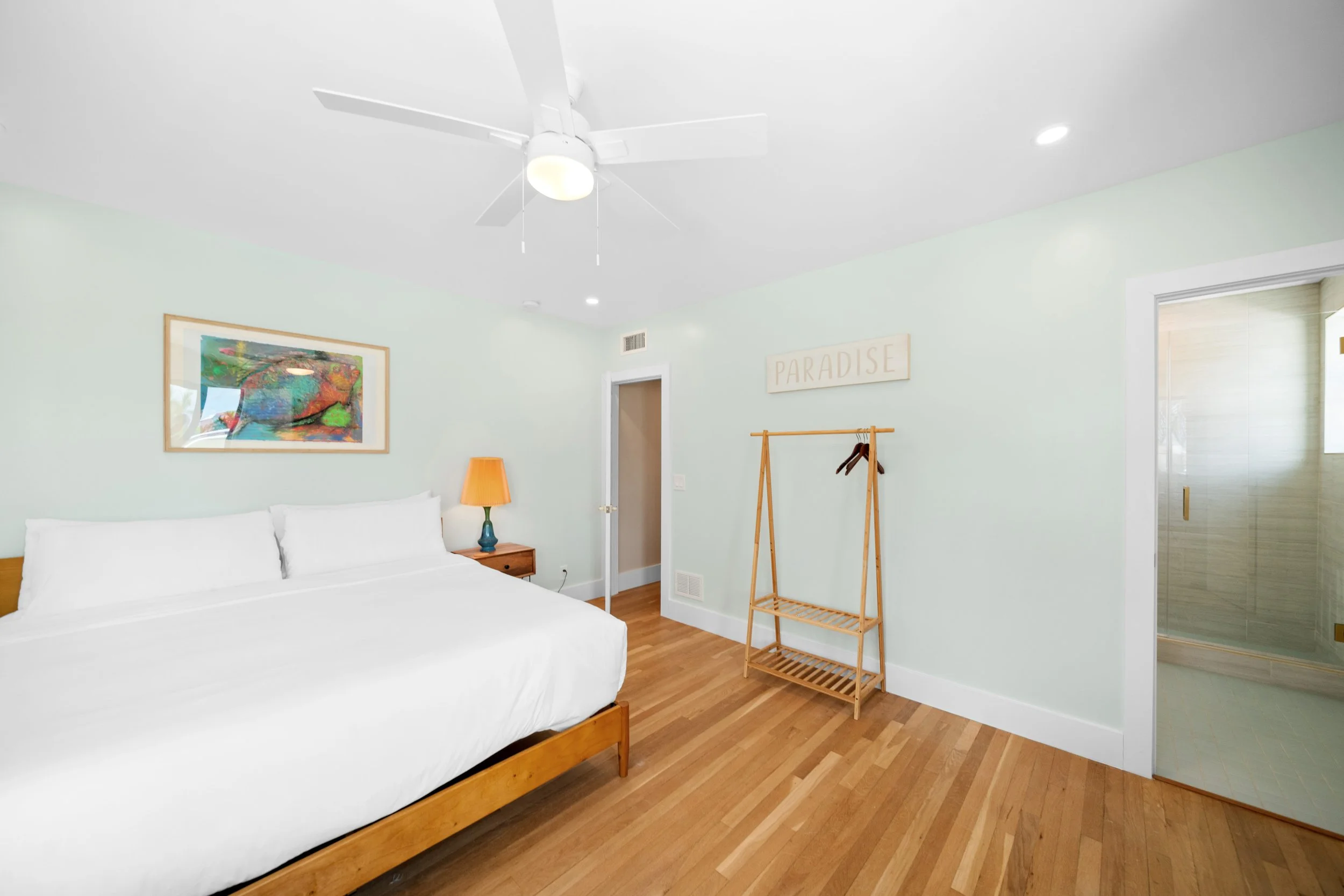 A bright bedroom with mint green walls, a white ceiling fan, a large bed with white bedding, a wooden nightstand with an orange lamp, a colorful framed painting, a wooden clothing rack, a sign that says "PARADISE," and a bathroom visible through an o