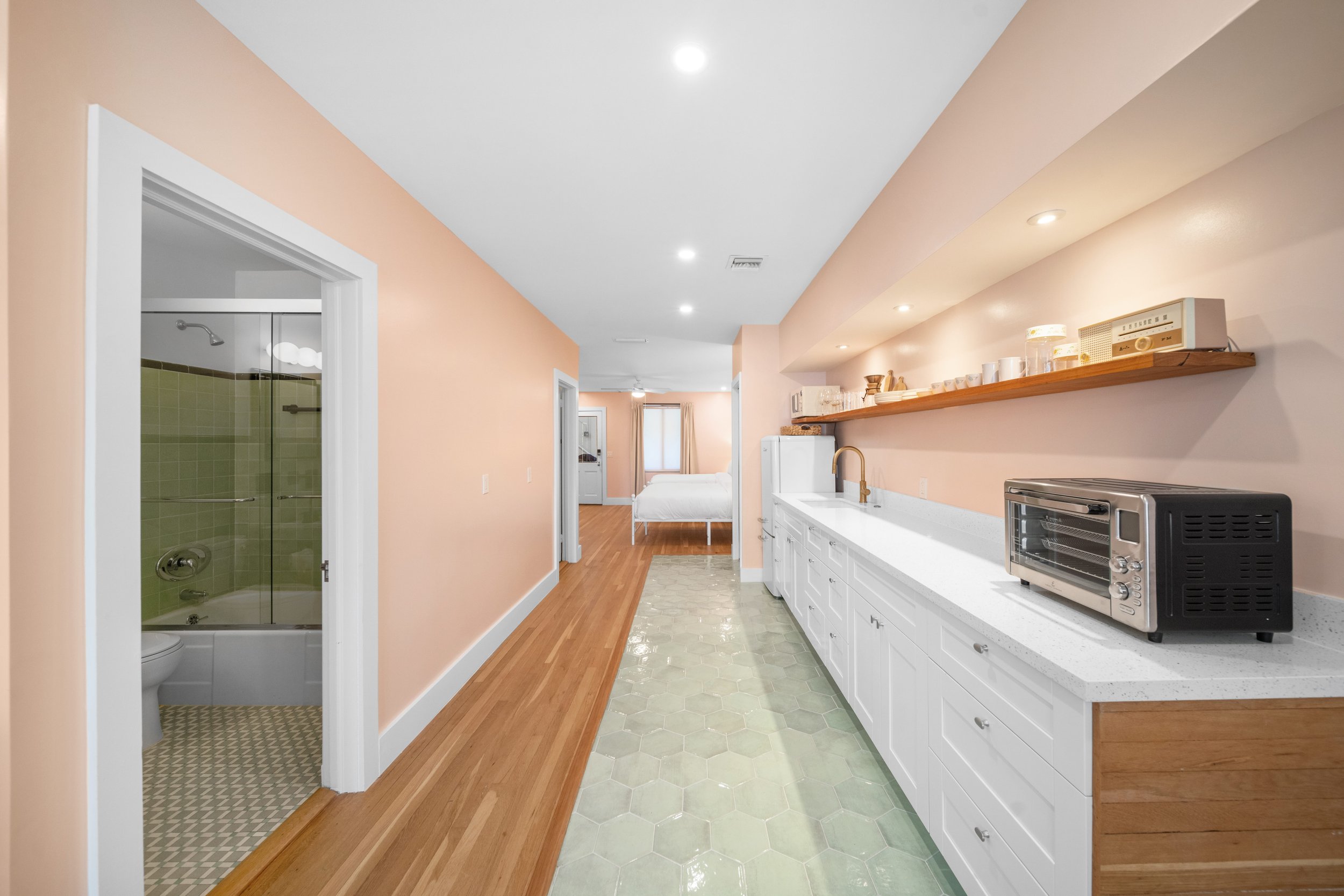 Kitchen with white cabinets, gold faucet, black toaster oven, open wooden shelf, peach walls, green hex tile floor, pink walls, doorway to bathroom with green tiled shower, and a view of bedroom with bed and window in the background.