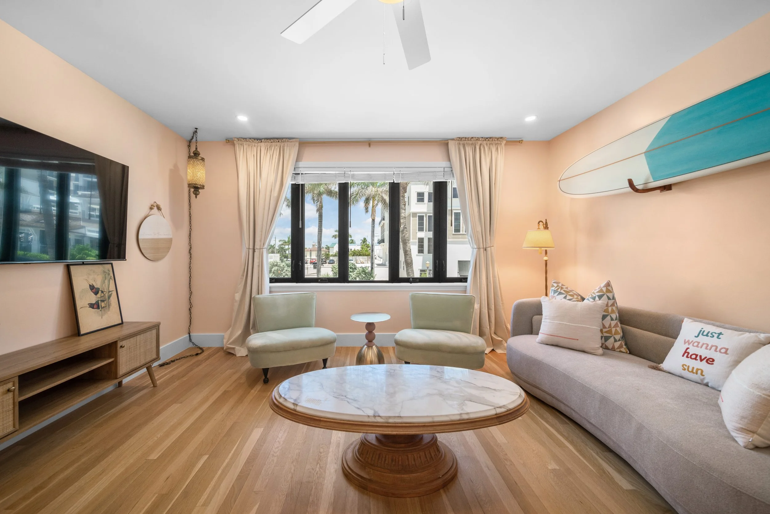 A living room with a large window showing palm trees outside. The room has a beige wall, a flat-screen TV on a wooden stand, two light green chairs, a grey sofa with decorative pillows, a marble-topped coffee table, and other decorative elements incl