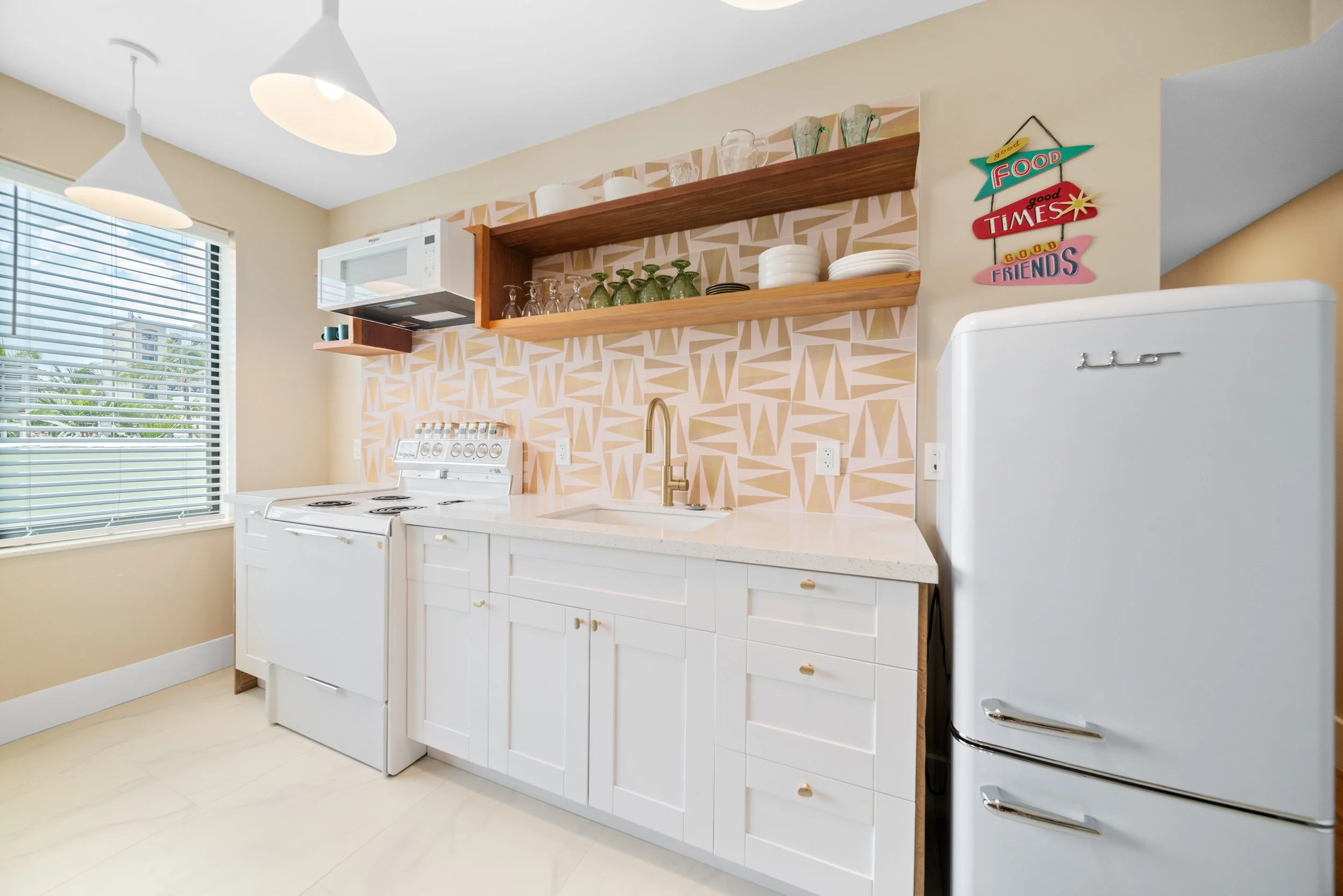 A modern kitchen with white cabinets, a white vintage refrigerator, a white stove, a white countertop with a sink and gold faucet, open wooden shelves with glassware and plates, a microwave, and a window with blinds. Bright lighting fixtures hang fro