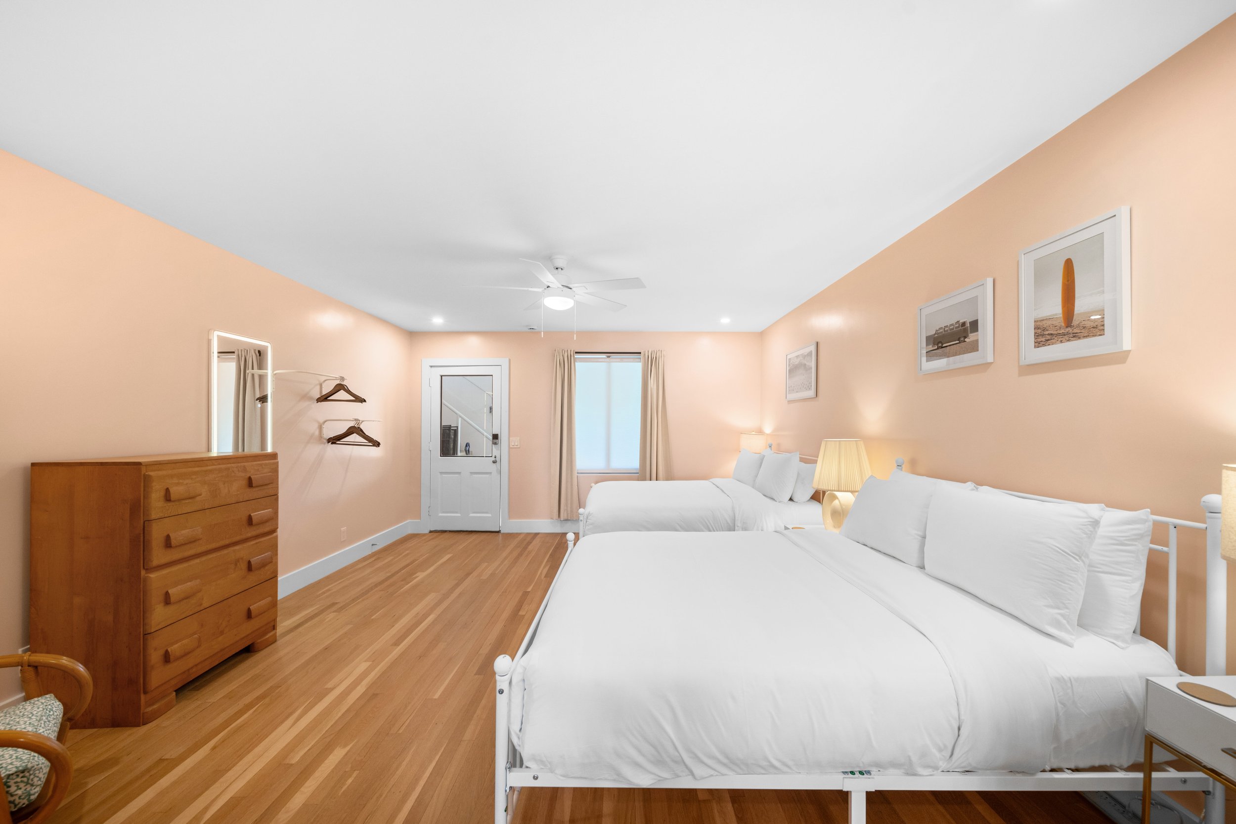 A bright hotel room with two white beds, wooden furniture, artwork on the walls, and a window with curtains.