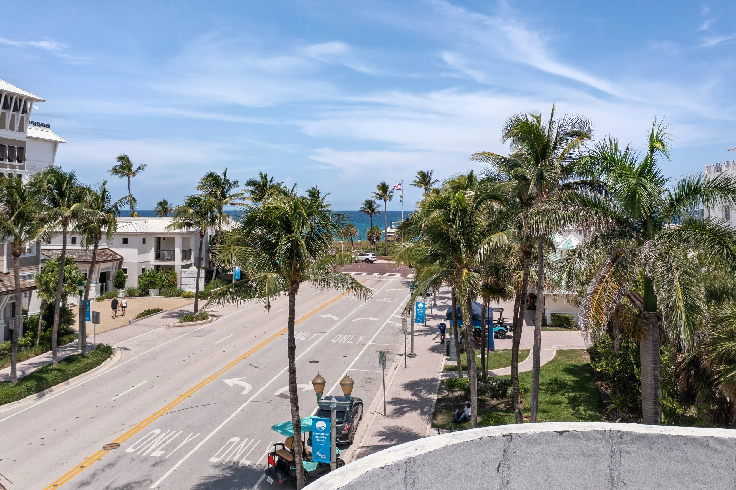 A sunny street scene near the beach with palm trees, white buildings, and the ocean visible in the background. There are a few people walking, cars parked, and a golf cart, with sky and clouds overhead.
