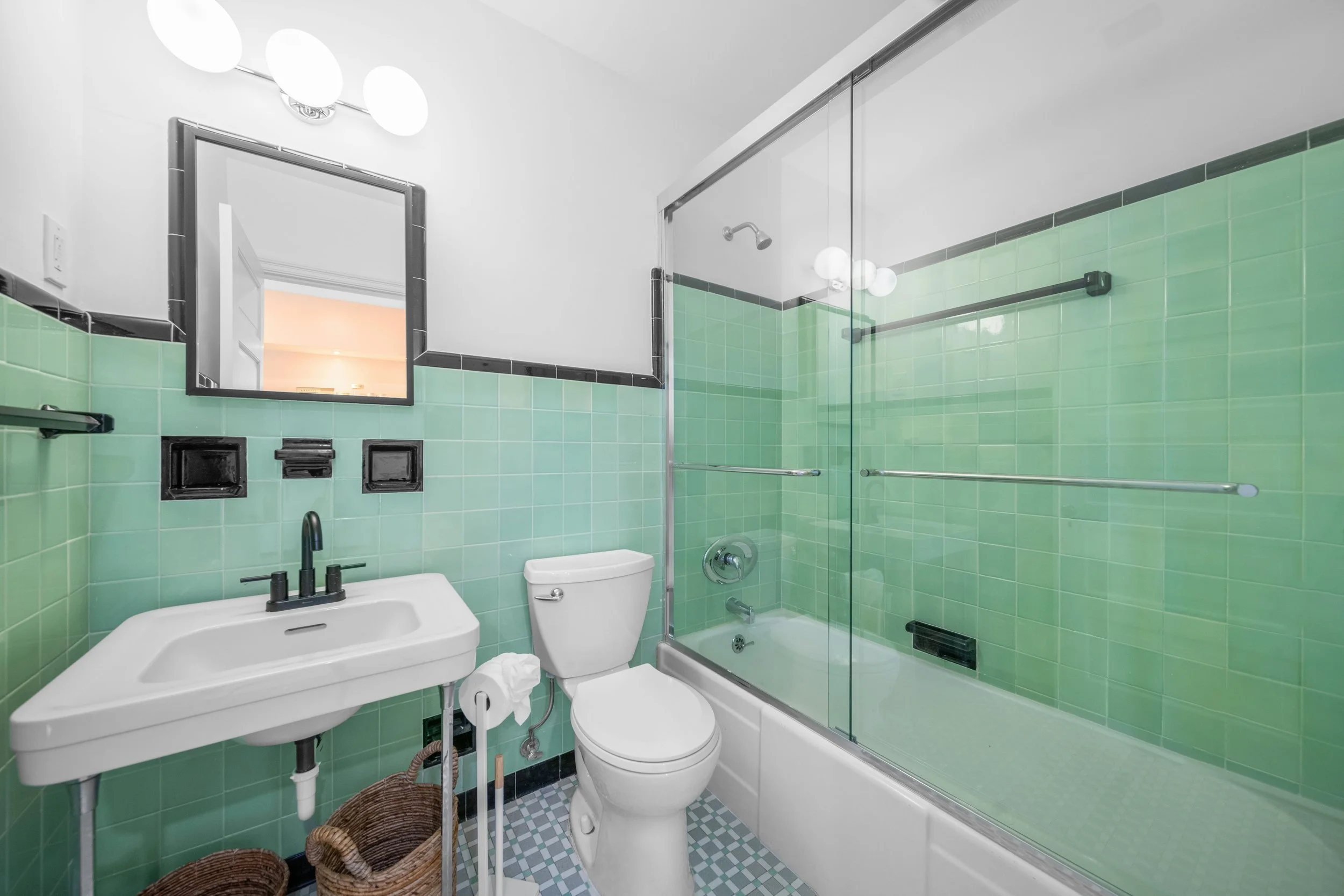 Bathroom with green tiled walls, a white toilet, a white sink, a mirror, and a glass-enclosed shower.