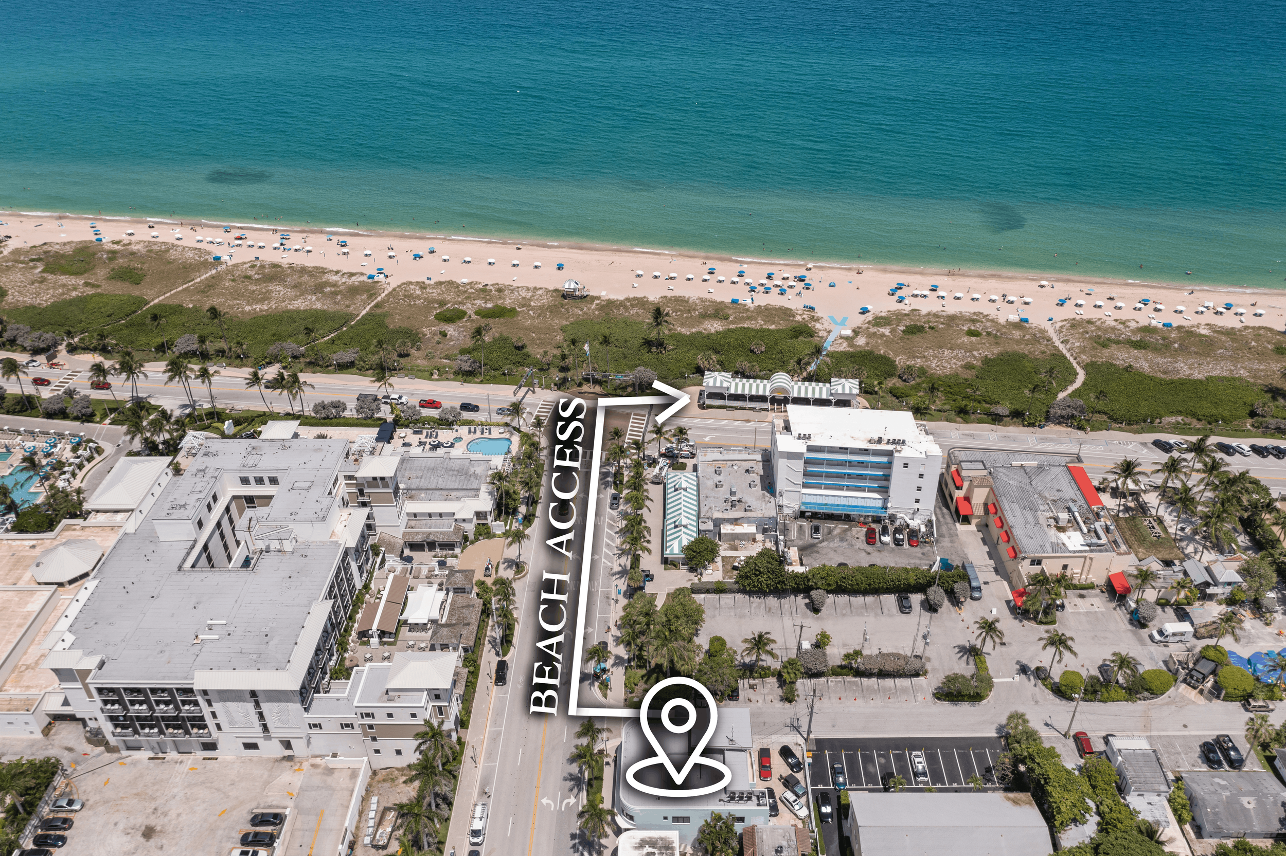 Aerial view of a beachside area showing a beach with umbrellas and umbrellas, buildings, parking lots, and a sign indicating 'Beach Access' with an arrow pointing toward the beach.