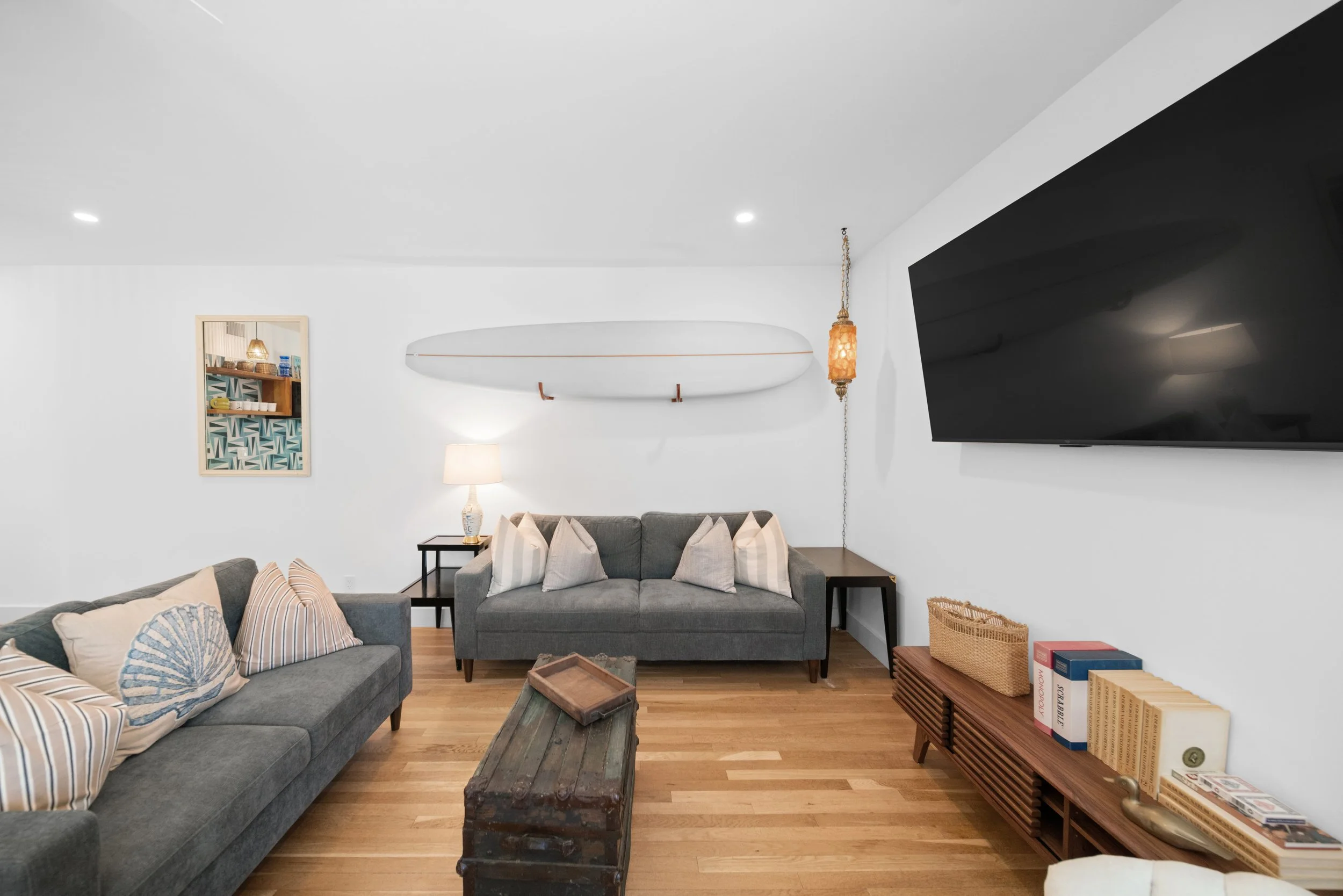 Living room with two gray sofas, wooden floor, wall-mounted TV, surfboard decor, bookshelf with books, lamp, and vintage chest as coffee table.
