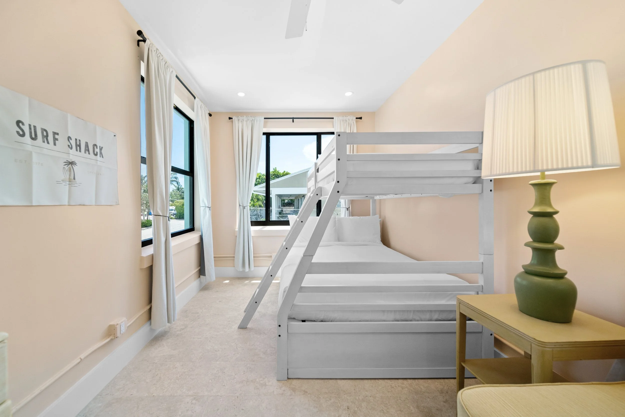 A bedroom with a white bunk bed, beige walls, cream carpet, a nightstand with a green lamp, and windows with white curtains showing a view of trees and a patio.