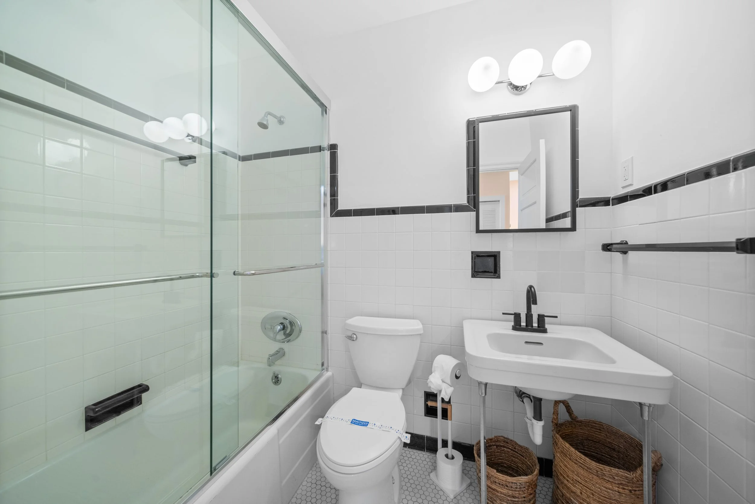 Small bathroom with a bathtub and shower, white toilet, wall-mounted mirror, black fixtures, and two woven baskets on the floor.