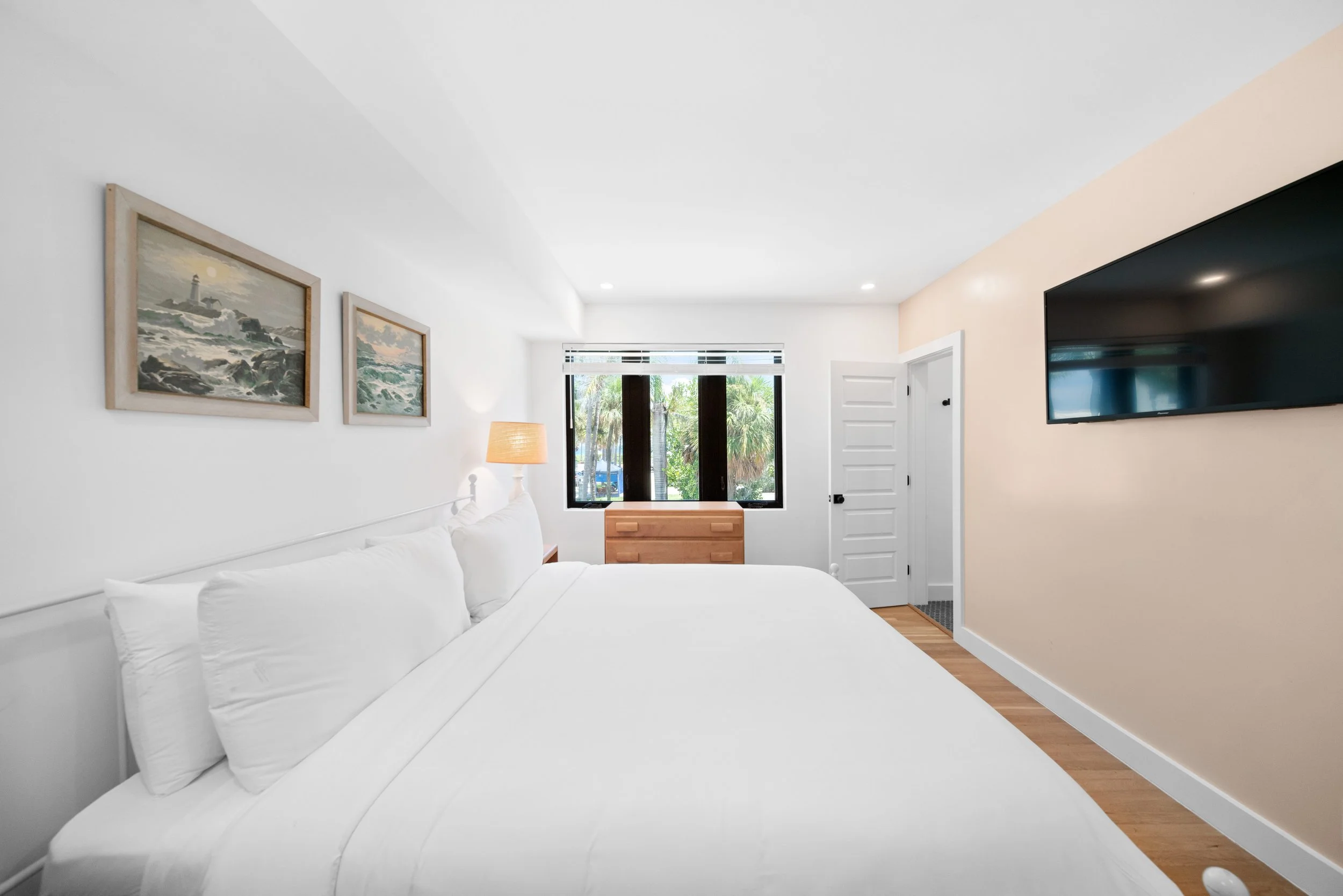 A bright hotel room with a large bed, white bedding, and pillows, a wooden nightstand with a lamp, framed artwork of ocean scenes on the wall, a window with greenery outside, a flat-screen TV mounted on a beige wall, and hardwood flooring.