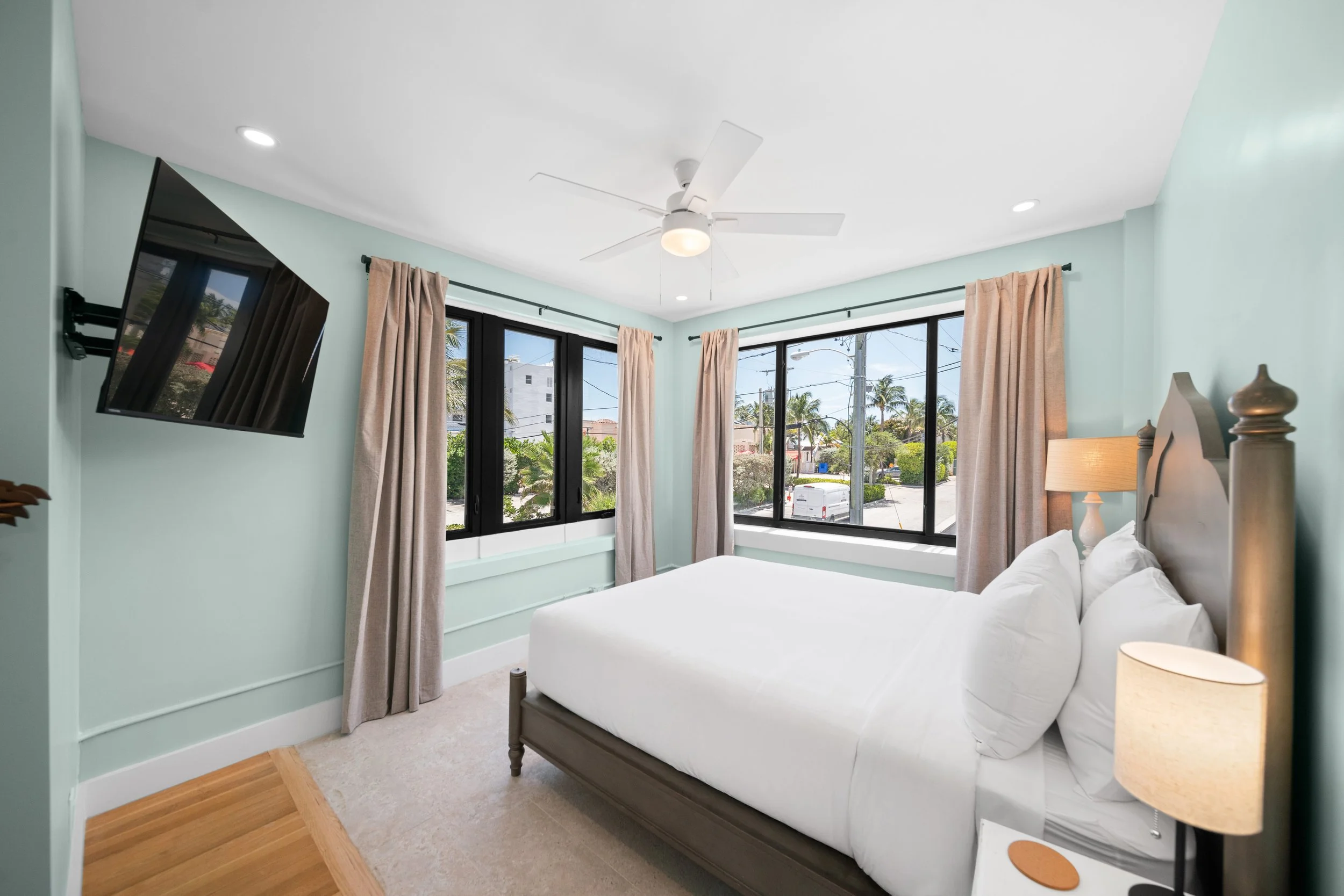 A bedroom with a bed, lamp, ceiling fan, large windows with curtains, and a wall-mounted TV.