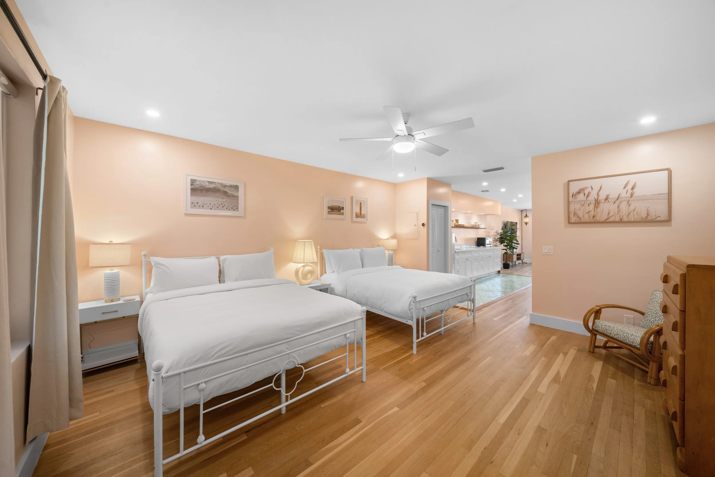 A spacious bedroom with two white beds, wooden floors, peach-colored walls, artwork above the beds, and a ceiling fan. In the background, there is a kitchen area with white cabinets and a small sitting area with a chair and potted plant.