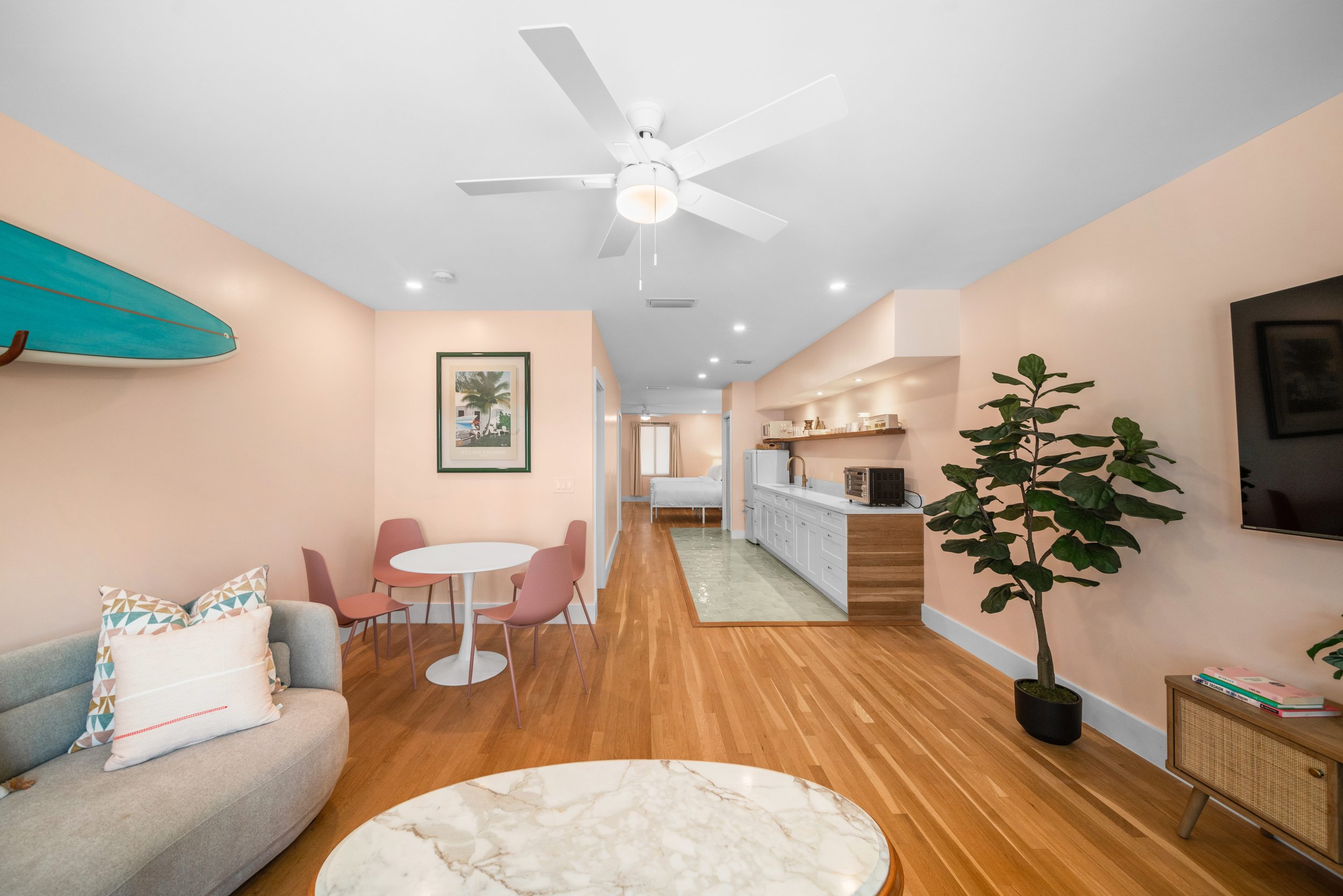 Open-concept living room with pastel walls, wooden flooring, a gray sofa with pillows, a white marble coffee table, pink dining chairs around a white table, a large potted plant, and a wall-mounted TV.
