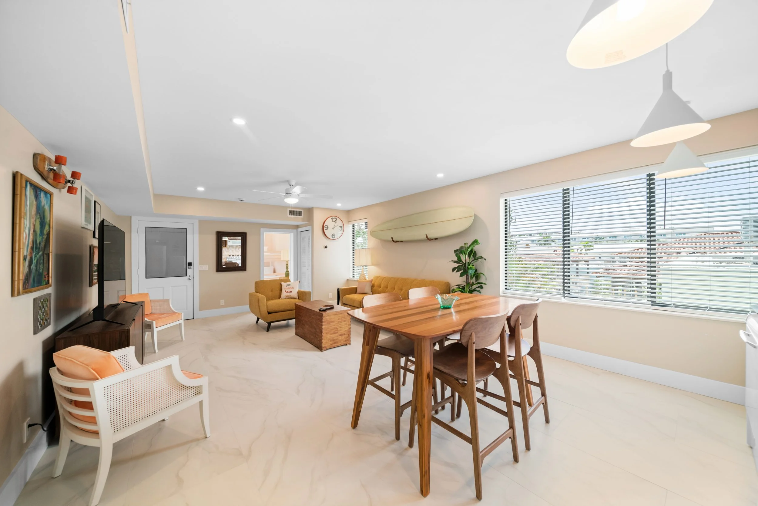 Bright living room and dining area with beige walls, large windows with blinds, tropical decor, mid-century modern furniture, and a surfboard on the wall.
