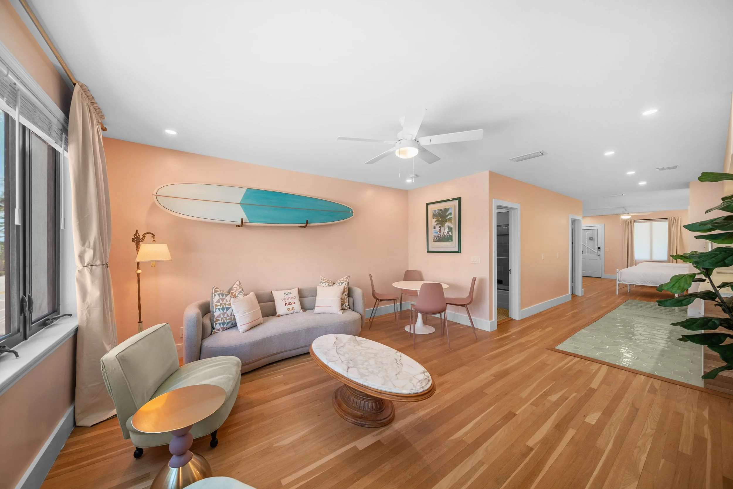 Living room and bedroom in a modern home with peach-colored walls, hardwood floors, a ceiling fan, a surfboard mounted on the wall, a sofa, an armchair, a marble coffee table, a dining table with four chairs, and a bed in the background.