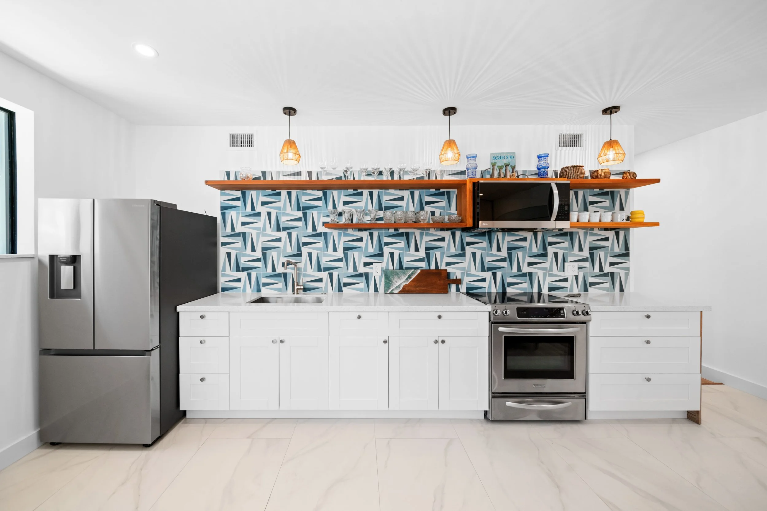 Modern kitchen with white cabinets, a geometric blue and gray tiled backsplash, stainless steel refrigerator and oven, open wooden shelves with glassware, and pendant lights.
