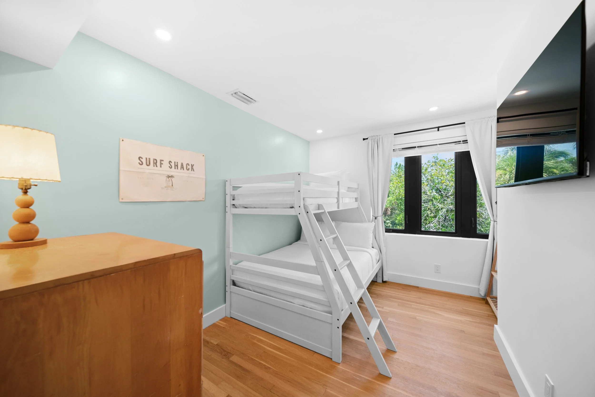 A bedroom with a white bunk bed near a window with white curtains and a view of green trees. There is a wooden dresser with a lamp on it and a wall sign that says "Surf Shack." A wall-mounted TV is on the right side of the image.