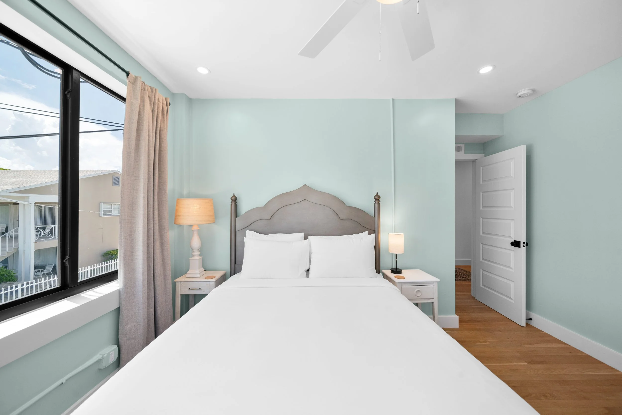 A bedroom with a large bed, white bedding, and a light wooden headboard. There are two bedside tables with lamps, one with a beige shade and the other with a black base. A window with beige curtains is on the left side, and an open white door is on t