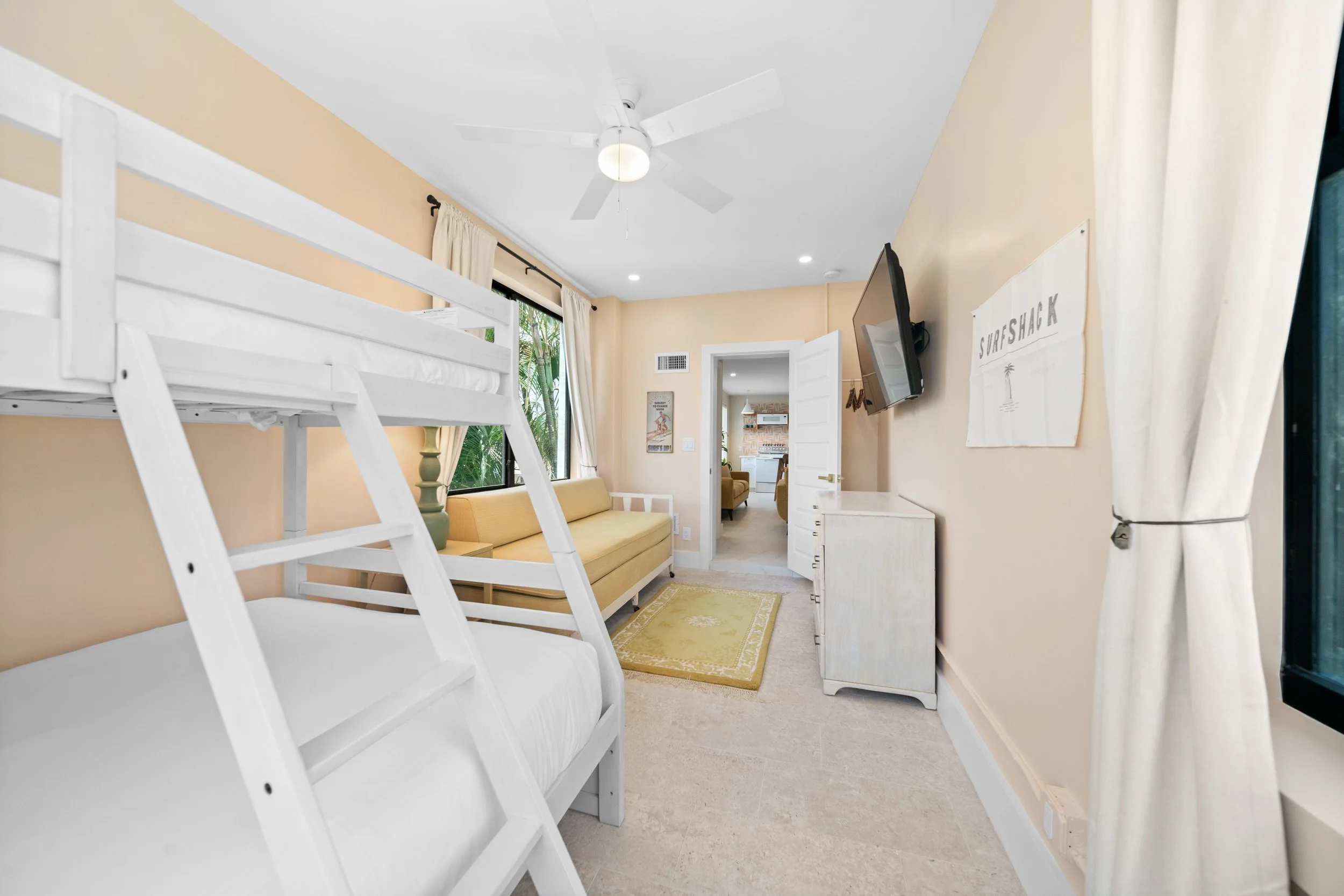 Interior of a bright bedroom with beige walls, a white bunk bed, a yellow sofa, a small white dresser, a wall-mounted TV, and a window with cream curtains.