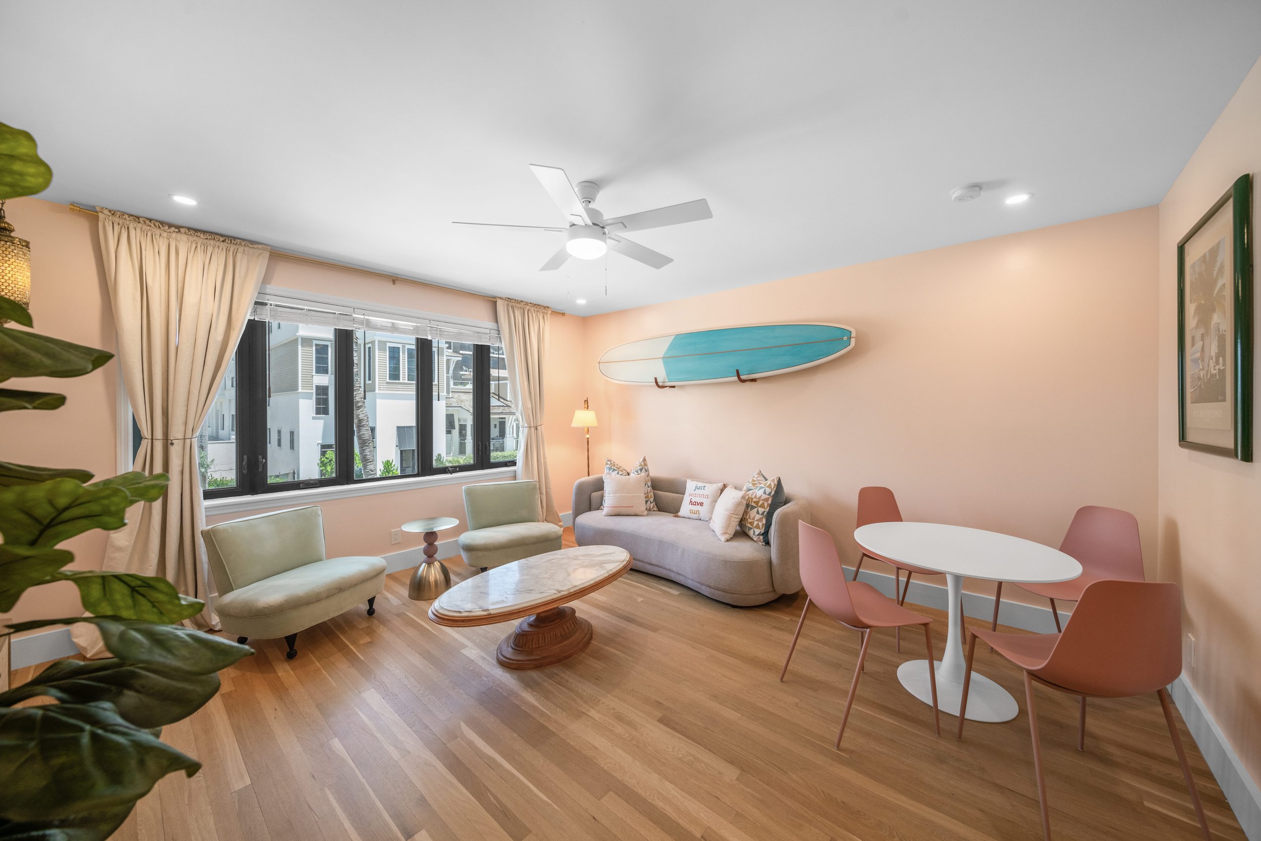 Living room with large window, cream curtains, beige walls, wooden floor, ceiling fan, white sofa with pillows, two light green chairs, a wooden coffee table, pink dining chairs around a white round table, surfboard on the wall, plant in the foregrou