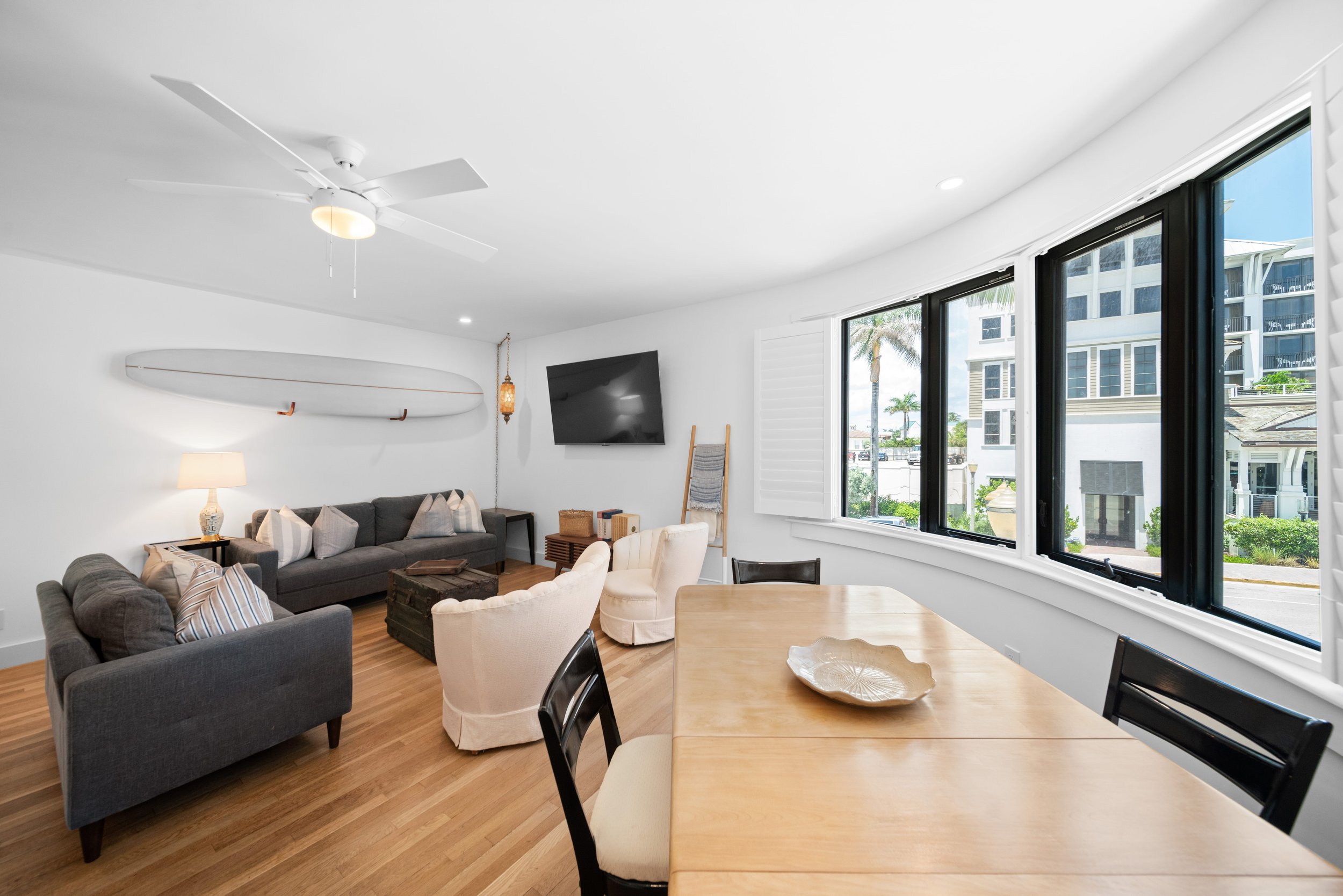 Bright, modern coastal living room with white walls, wooden floors, a ceiling fan, a wall-mounted flat screen TV, large windows with black frames, and furniture including gray and white sofas, chairs, and a dining table.