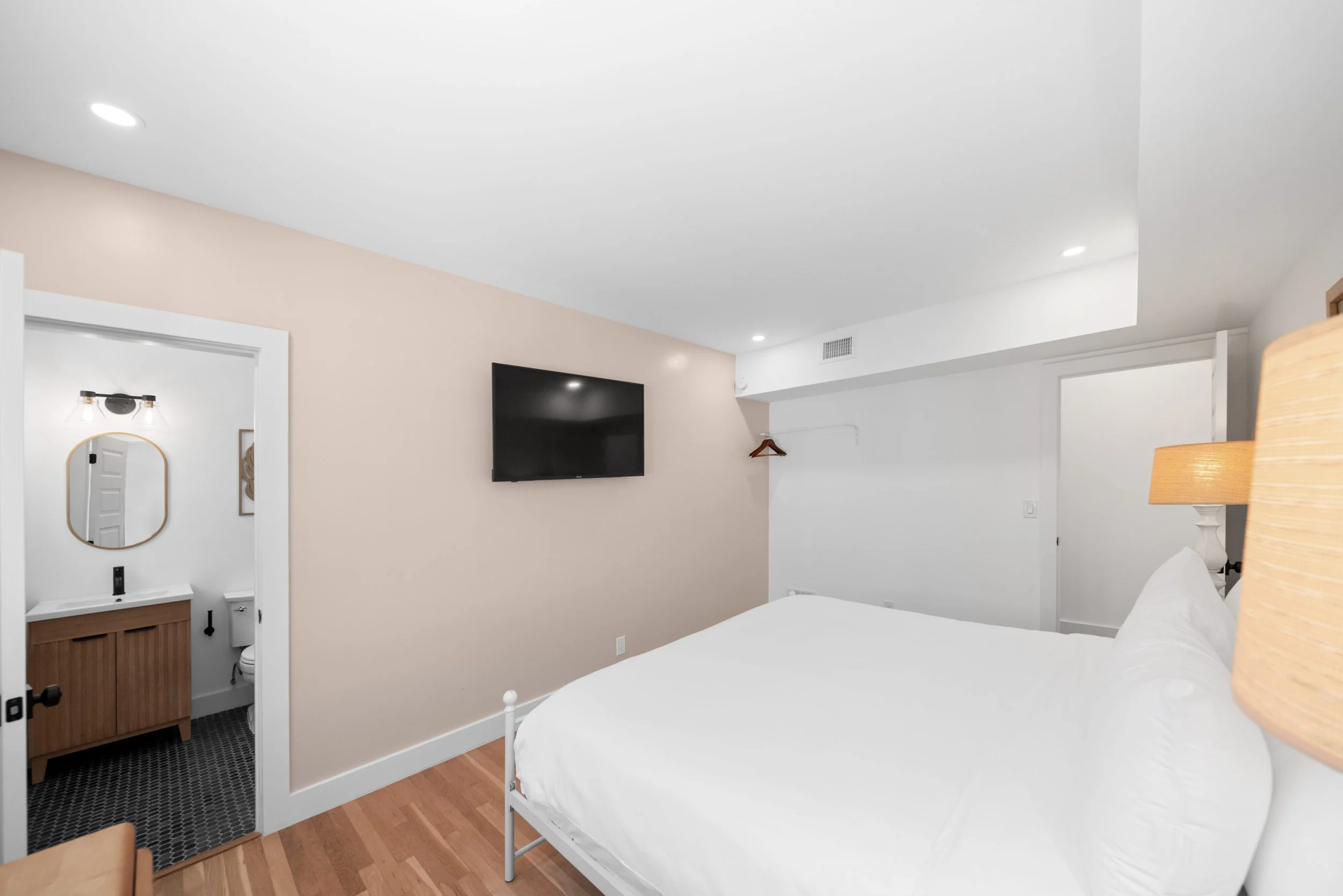 A bedroom with a white bed, wooden nightstand, wall-mounted TV, beige accent wall, and a small adjoining bathroom visible through an open door.