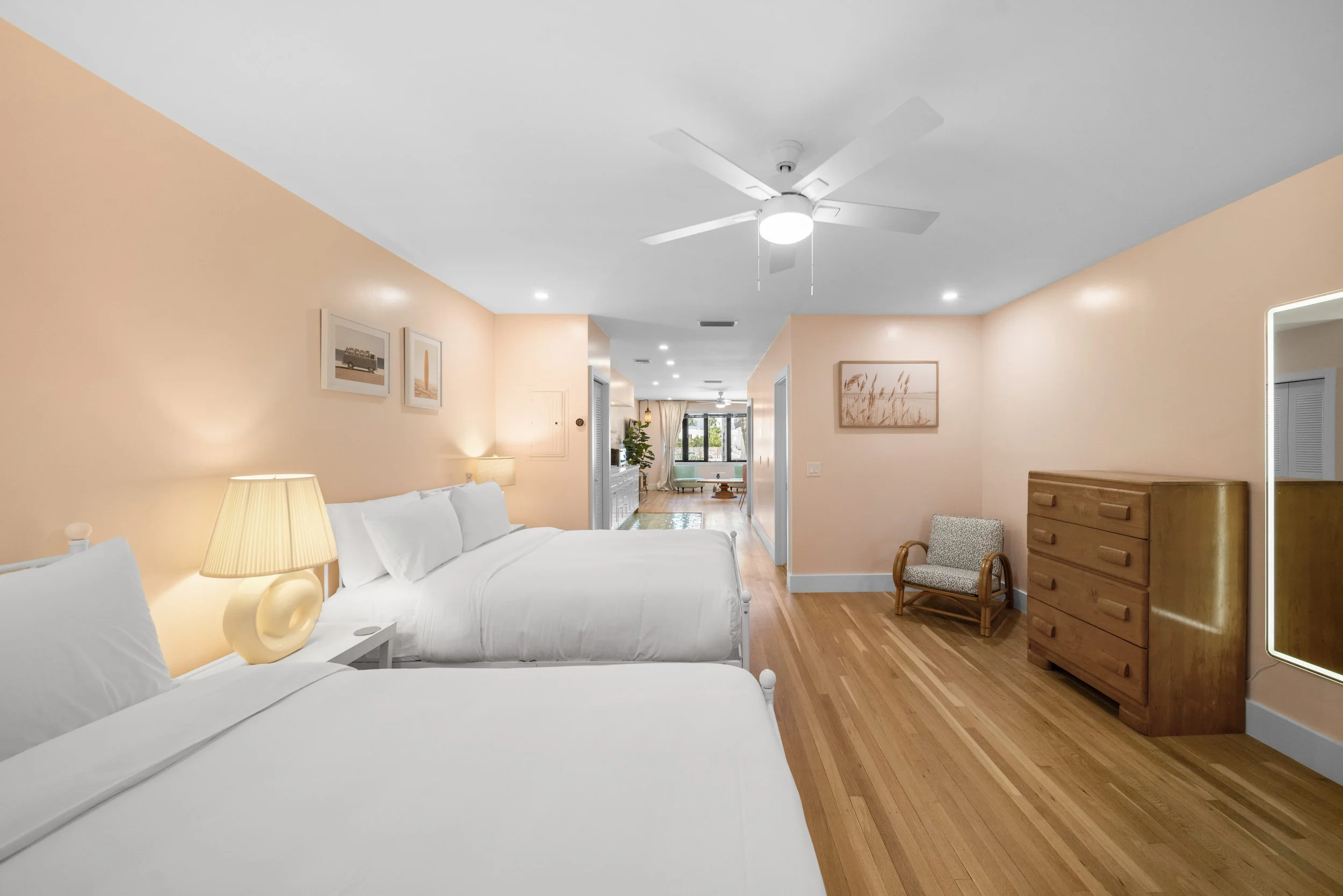 Hotel room with two beds, wooden dresser, armchair, framed artwork, and a view into living area with windows and furniture.
