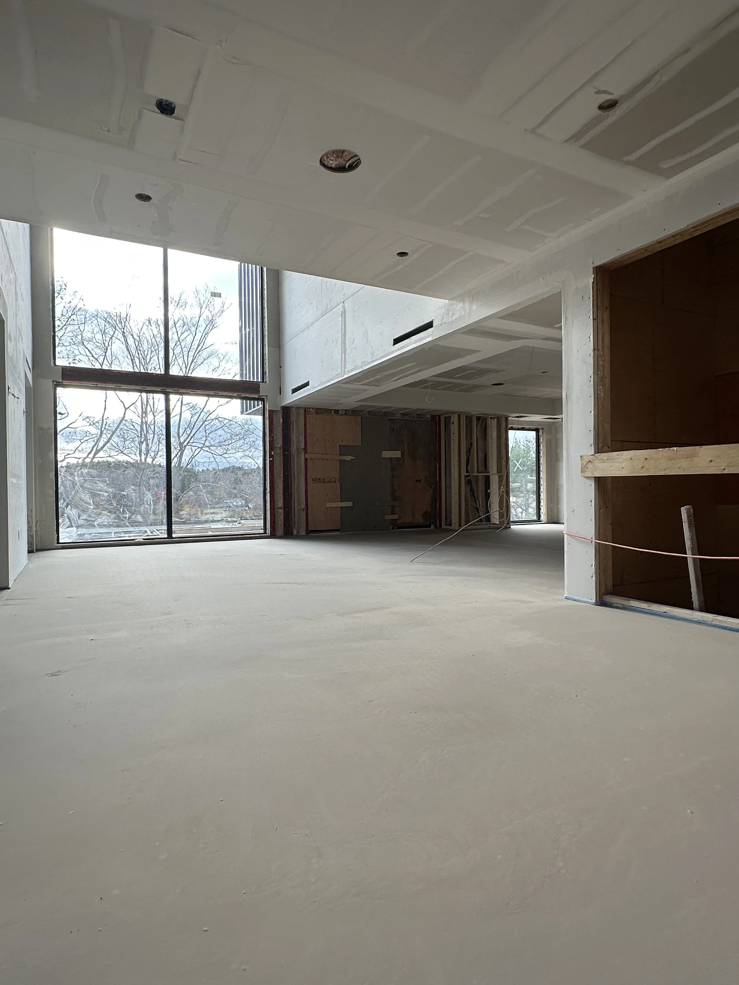 An unfinished room in a building under construction, with large window showing trees outside, drywall in progress, and open wall spaces.