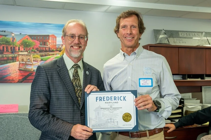 Two men smiling and holding a Frederick, Maryland certificate of memorials in an office with a colorful painting of a park in the background.