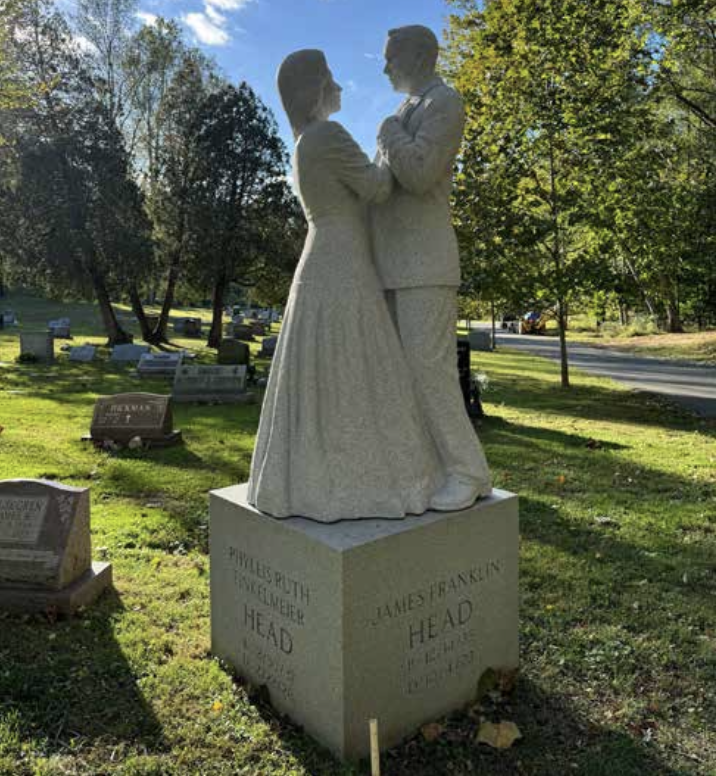 A white marble statue of a bride and groom facing each other, holding hands, in a cemetery with trees and headstones in the background.