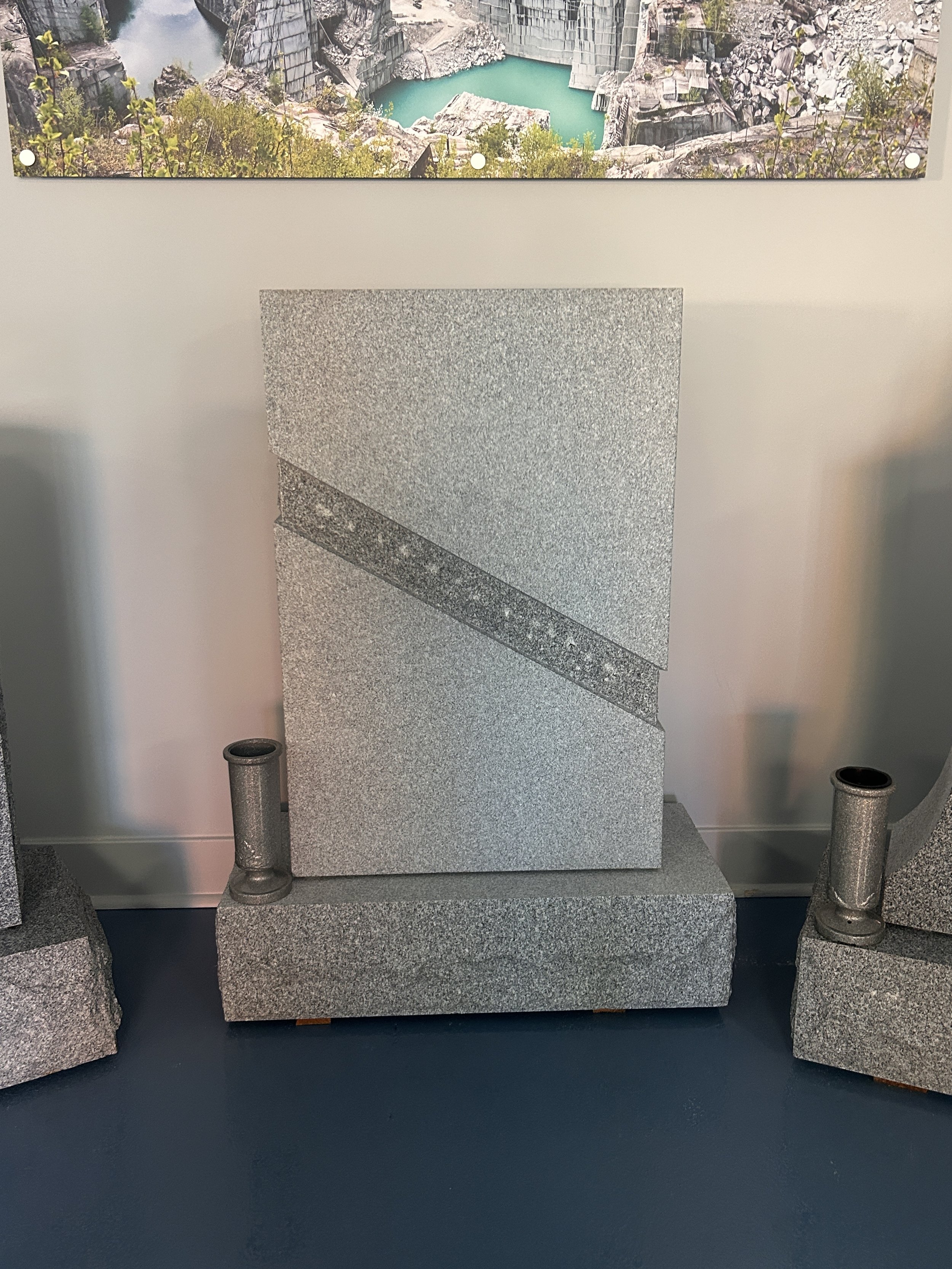 Display of a granite monument structure with four cylindrical granite bases, two at the front and two at the back, surrounding a central upright granite panel with a textured and veined surface. The background is a plain wall, and the floor is painted dark.