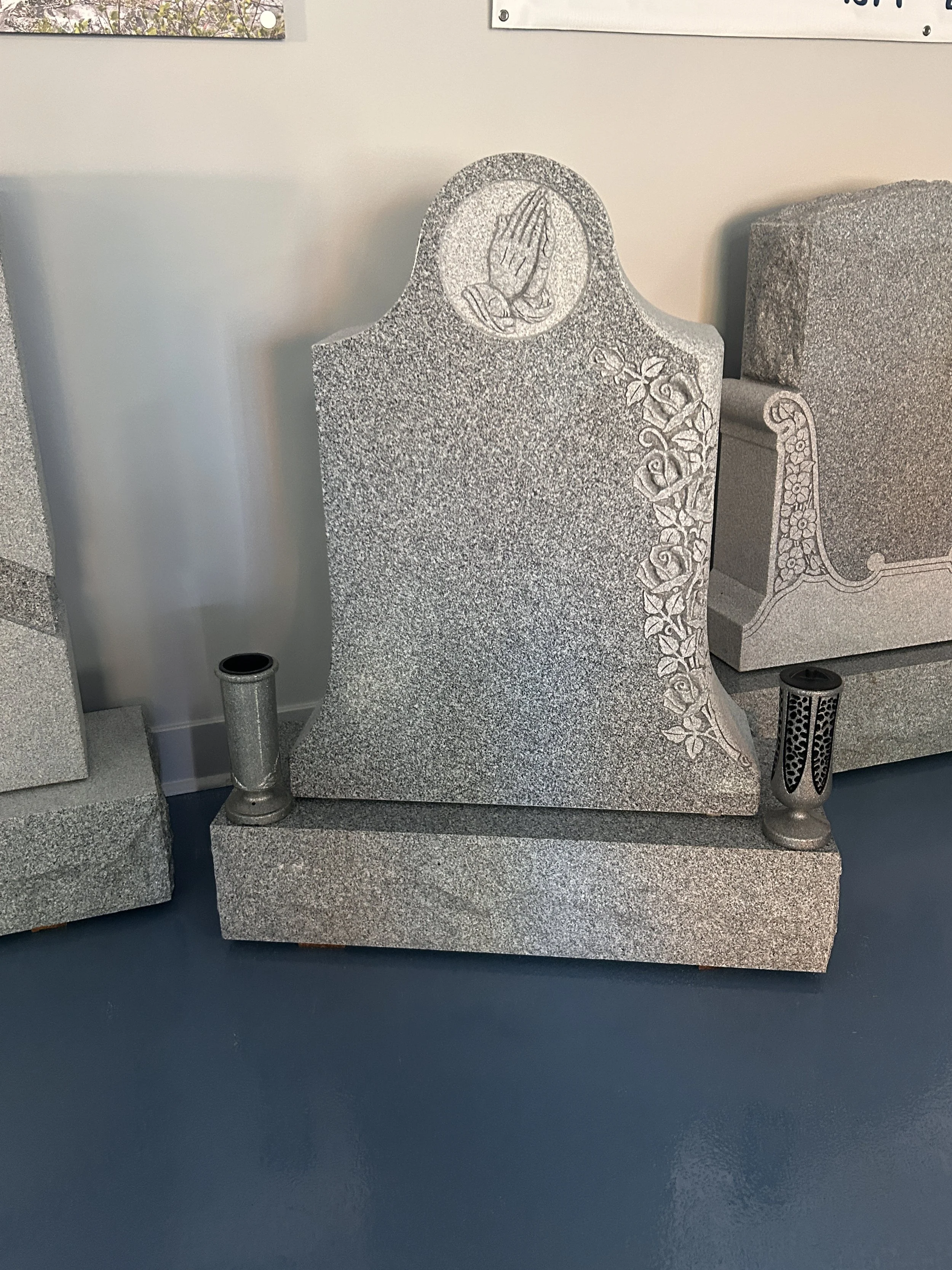 Gray granite headstone with engraved praying hands and a floral decorative border, displayed in a museum or memorial setting.