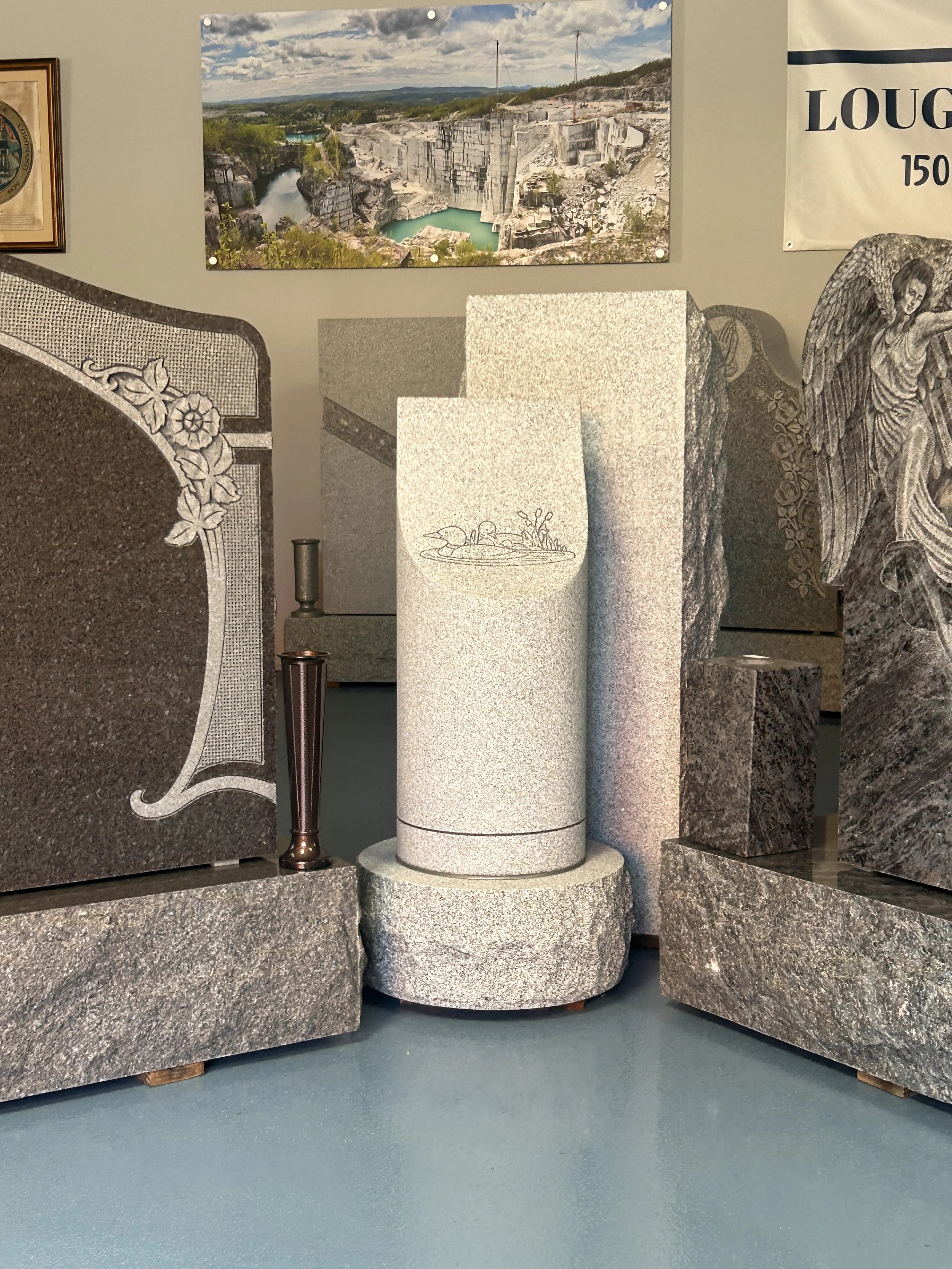 A selection of gravestones made from different types of stone, with a polished granite headstone on the left, a white granite cylindrical memorial in the center, and a dark polished stone with angel engraving on the right