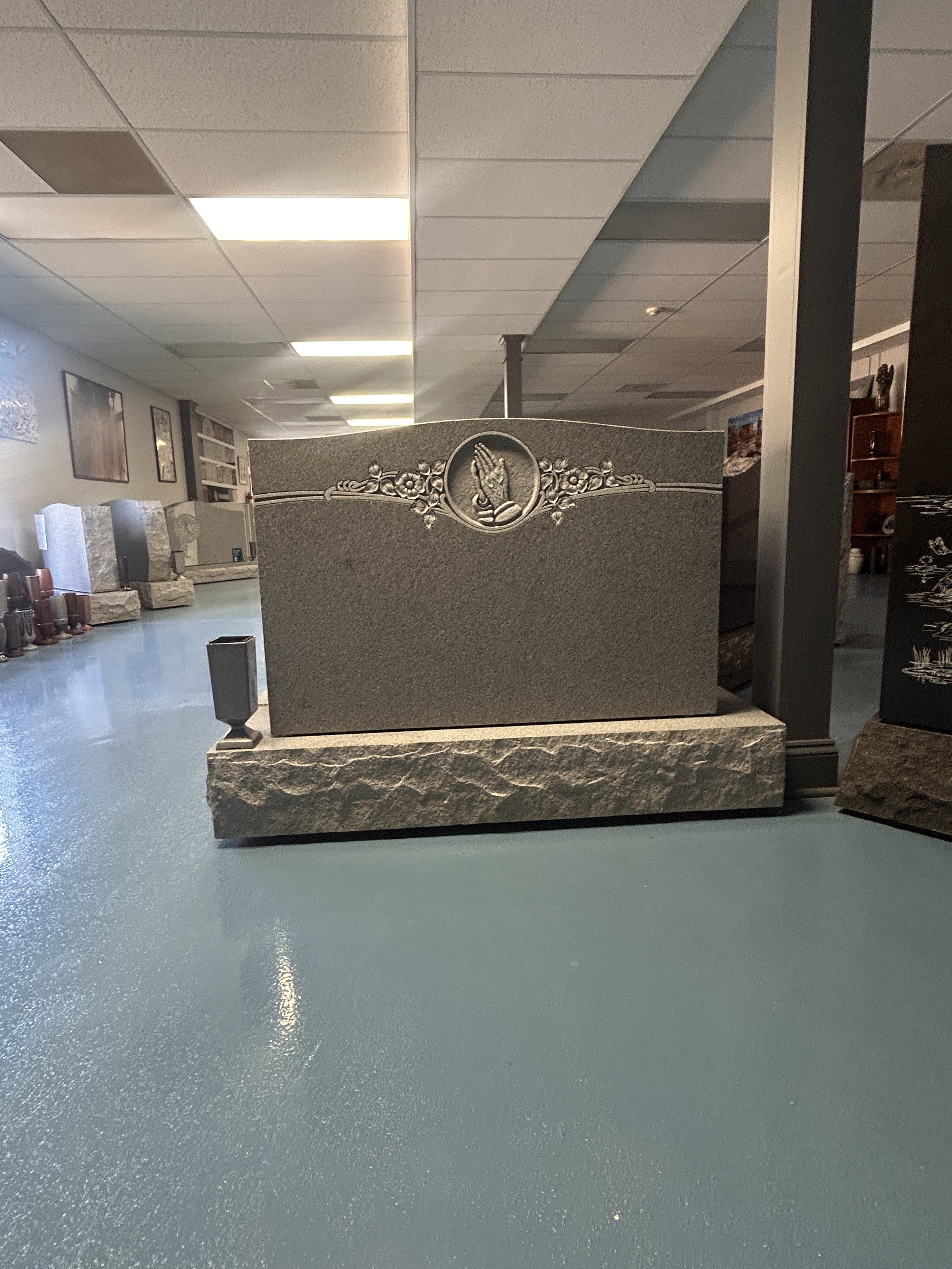 A granite tombstone with a carved praying hands symbol, placed on a flat base