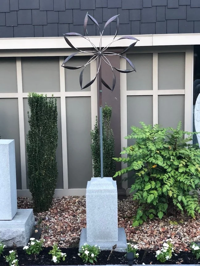 Metal sculpture of a geometric flower on a curved stem, mounted on a stone base, with green shrubs and a gray wooden fence in the background.