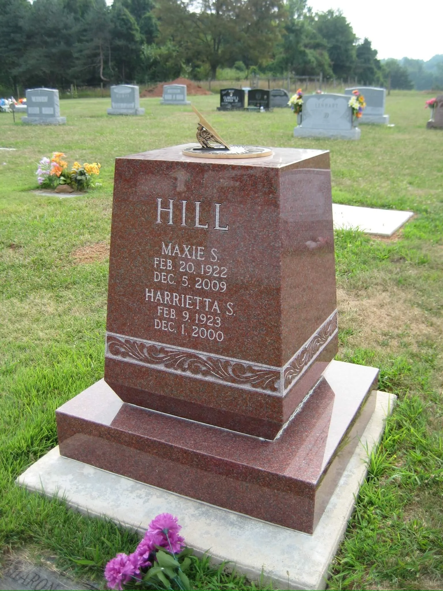 A red granite gravestone with an engraving of two birds and floral patterns at the bottom. The gravestone contains the names Maxie S. Hill and Harrietta S. Hill, with their birth and death dates. There are purple flowers planted at the base, and in the background, several other gravestones with flowers and decorations are visible in a green cemetery setting with trees.
