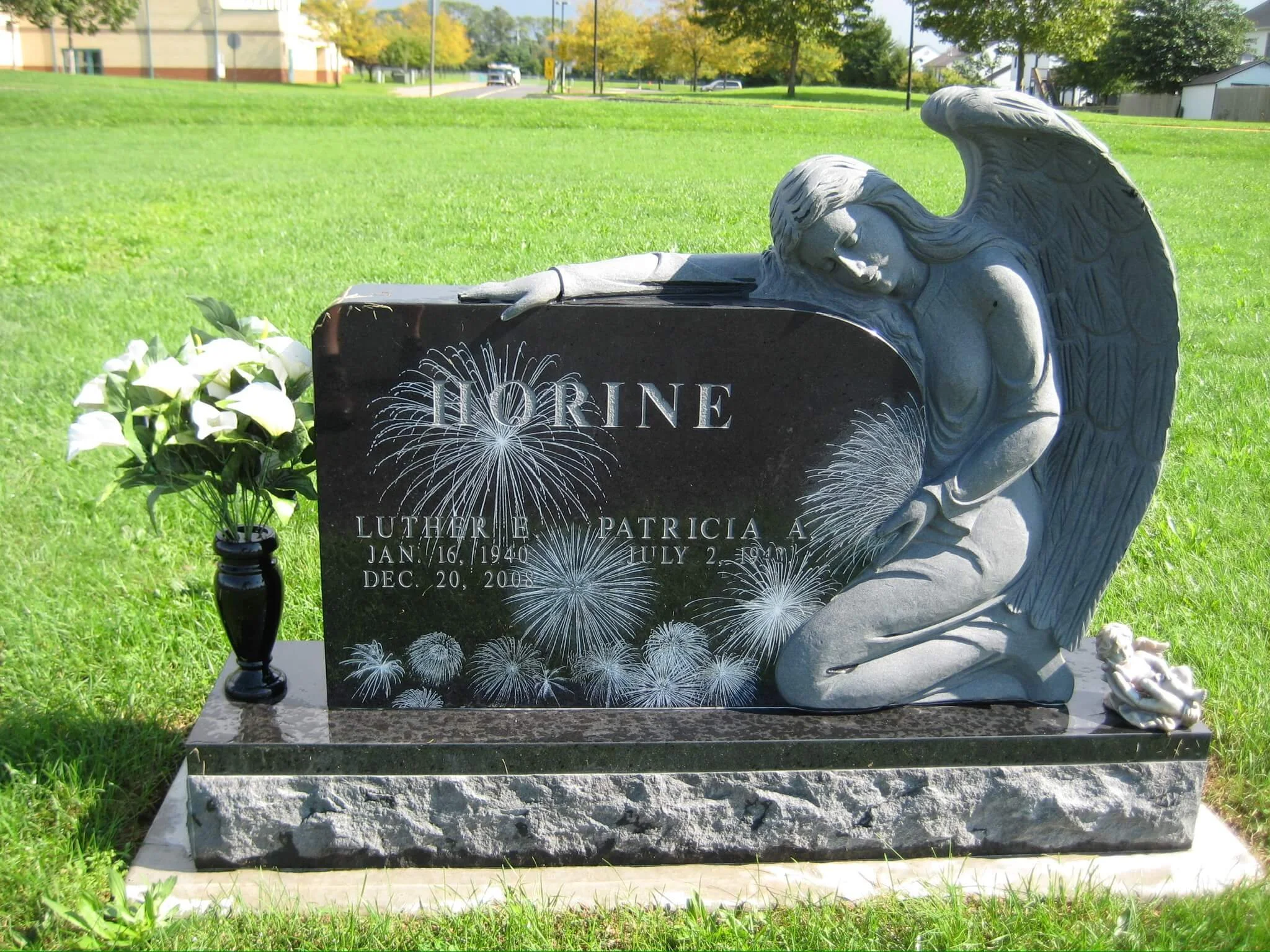 A gravestone with a praying angel sculpture lying on top of it. The gravestone has the name Loreine Patricia, with dates of birth January 16, 1940, and death December 20, 2008. There are white flowers and a small black lamp placed beside it in a grassy cemetery.