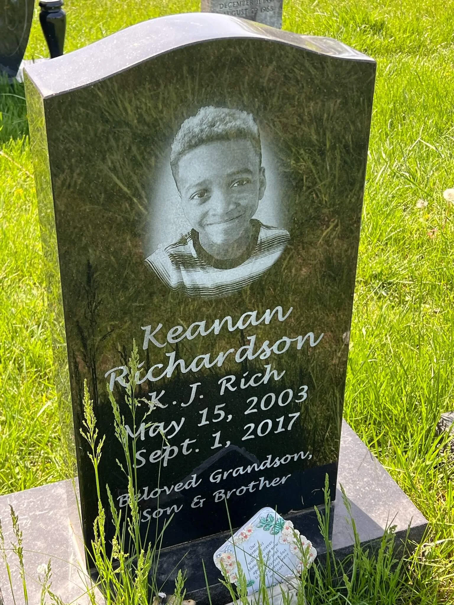 A black granite headstone with an engraved photo of a young boy, commemorating Keanan Richardson and listing his birth date as May 15, 2003, and his death date as September 1, 2017, with surrounding green grass.