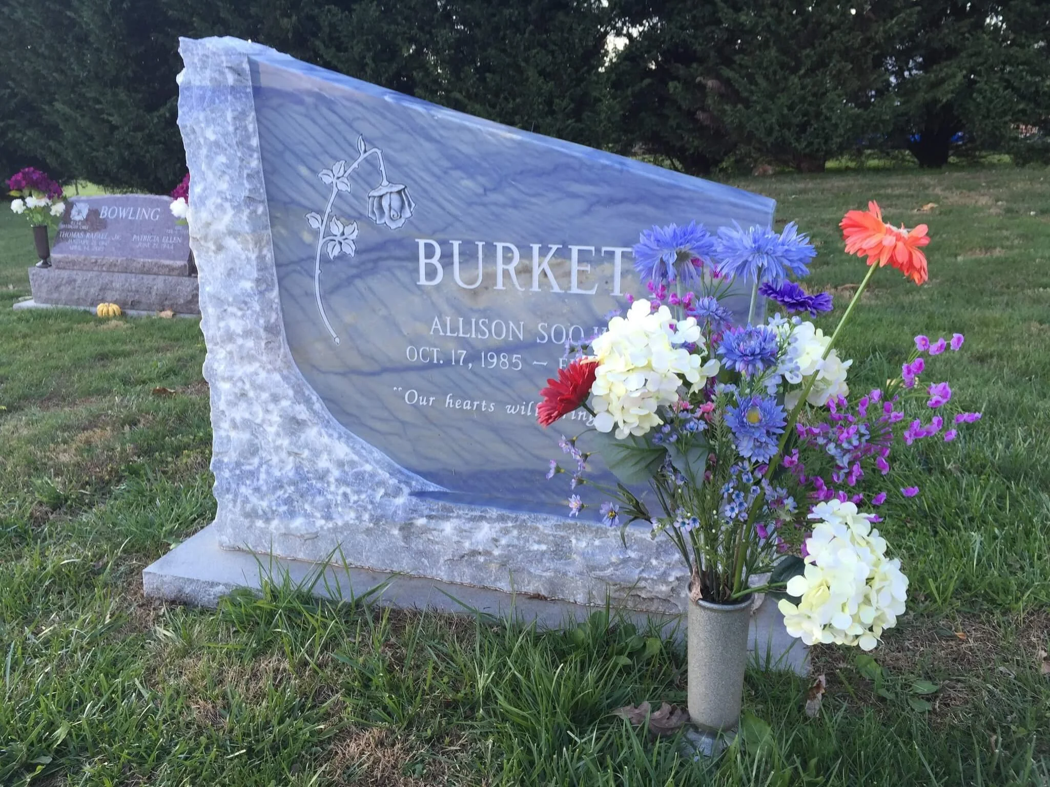 Gravestone with a flower arrangement in front, surrounded by grass and trees.
