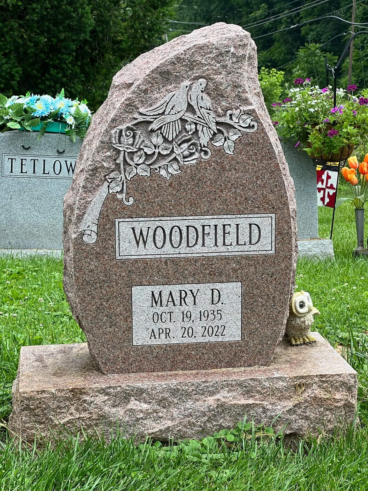 A gravestone with engraved details and a carved depiction of two birds on a branch. The gravestone reads 'Woodfield' and commemorates Mary D. who was born on October 19, 1935, and passed away on April 20, 2022. There are colorful flower arrangements and a small owl figurine nearby, with green grass and trees in the background.