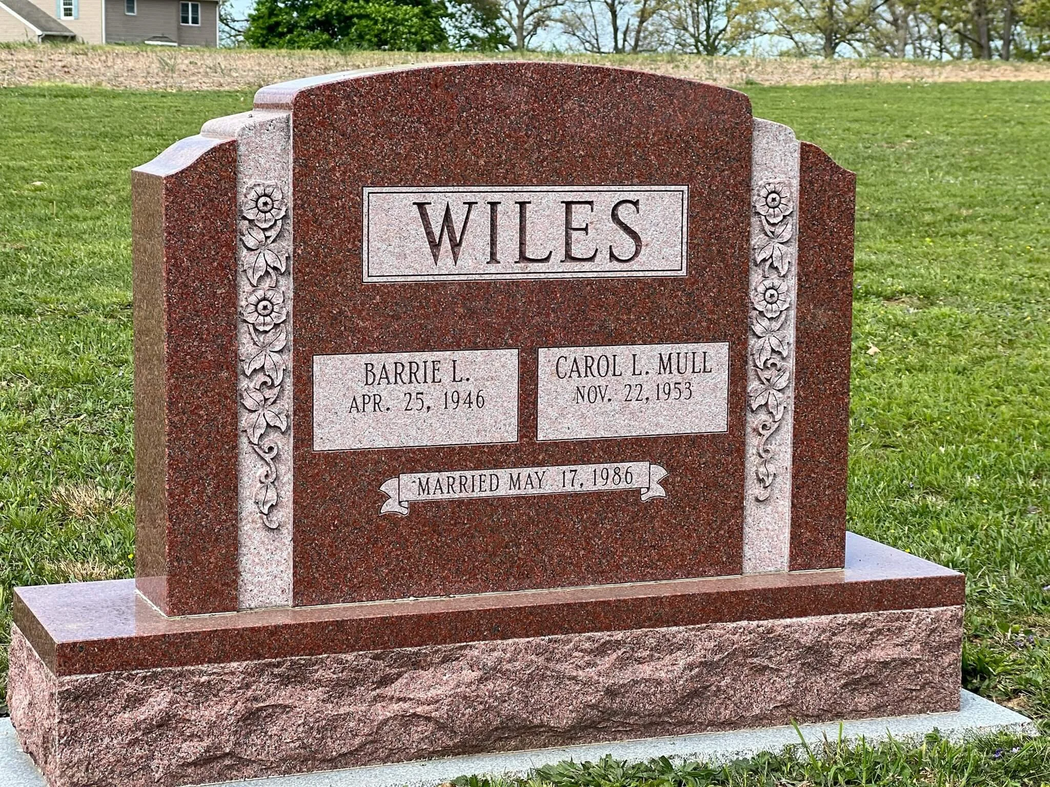 A red and gray granite tombstone with names and birthdates, surrounded by grass and trees in a cemetery.