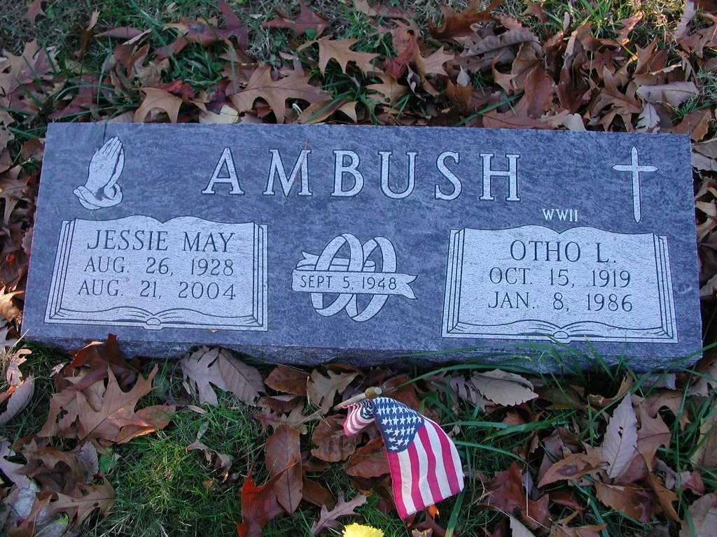 Gravestone with the surname 'Ambush,' engraved with the names Jessie May and Otho L., including birth and death dates, an open book symbol, a ribbon with a date, a praying hands symbol, and a cross.