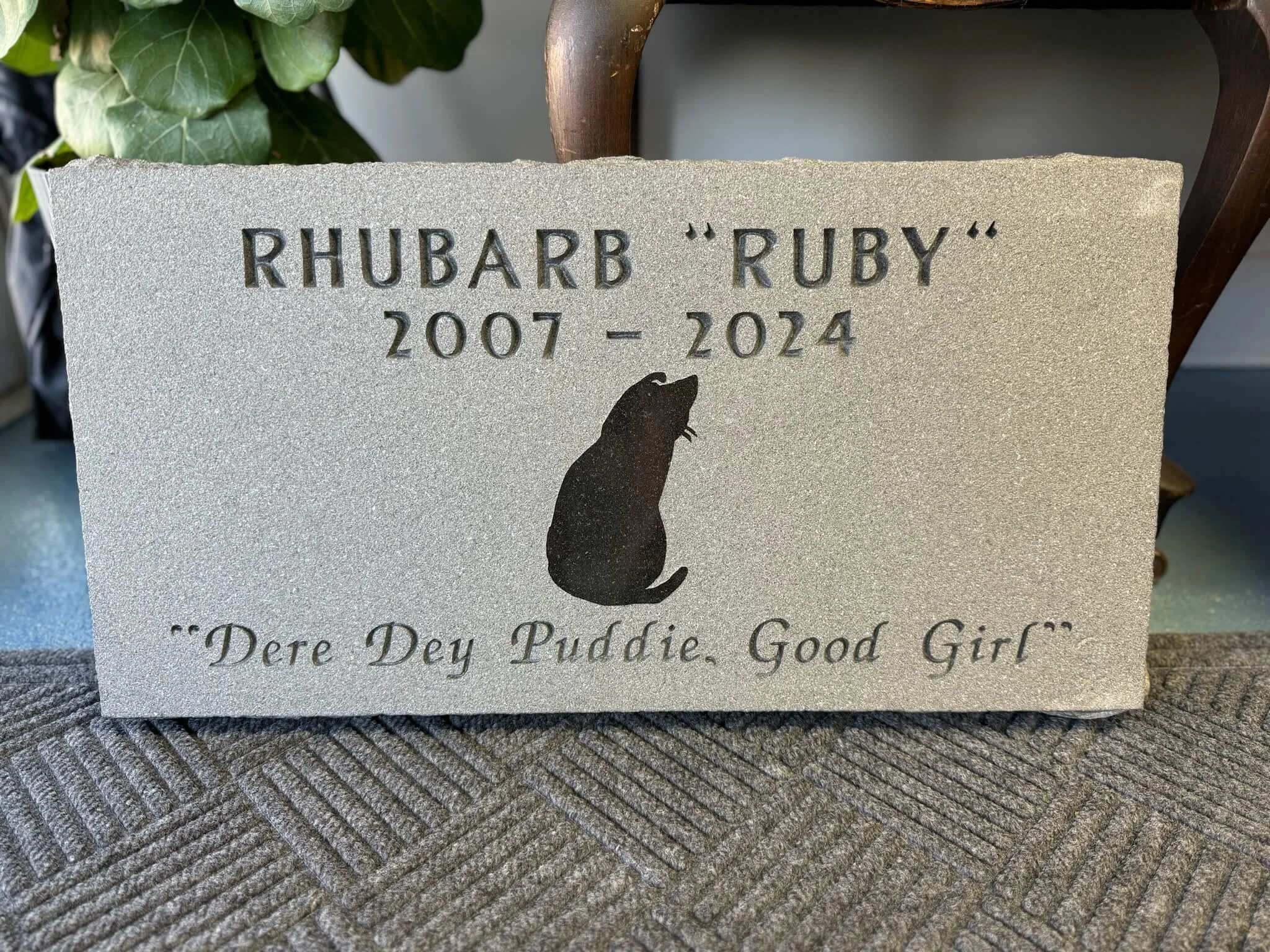A gravestone with the name 'Rhubarb 'Ruby,' her birth year 2007, and passing year 2024. The gravestone features a black silhouette of a hen and the quote, 'Dere Dey Puddie, Good Girl.'